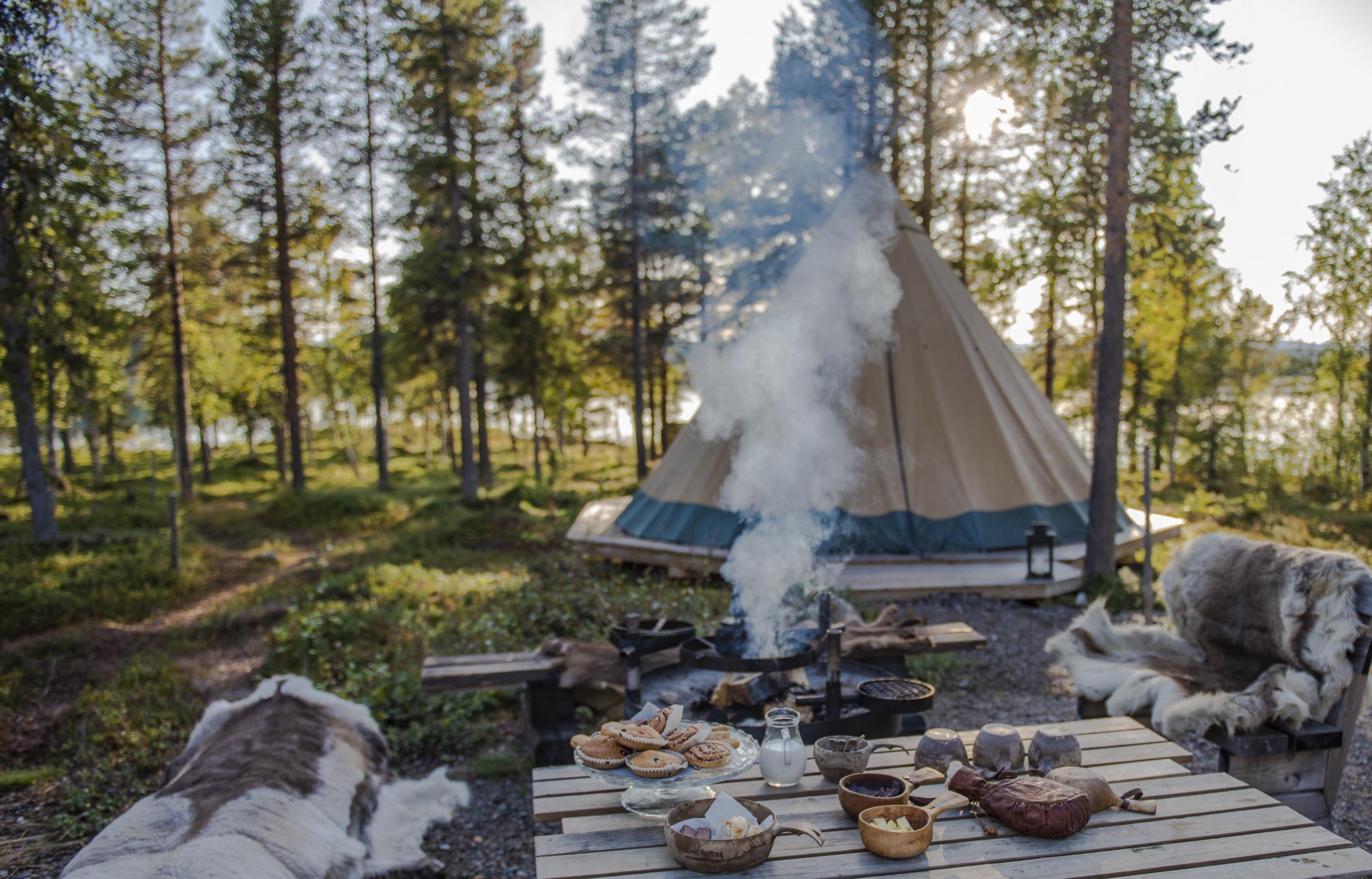 Banken bedekt met rendierhuiden naast een tafel met kopjes en schalen en een bord met broodjes. Een koffiepot wordt boven een kampvuur geplaatst. Een lavvu-tent op de achtergrond en je krijgt een glimp van een meer tussen de bomen.