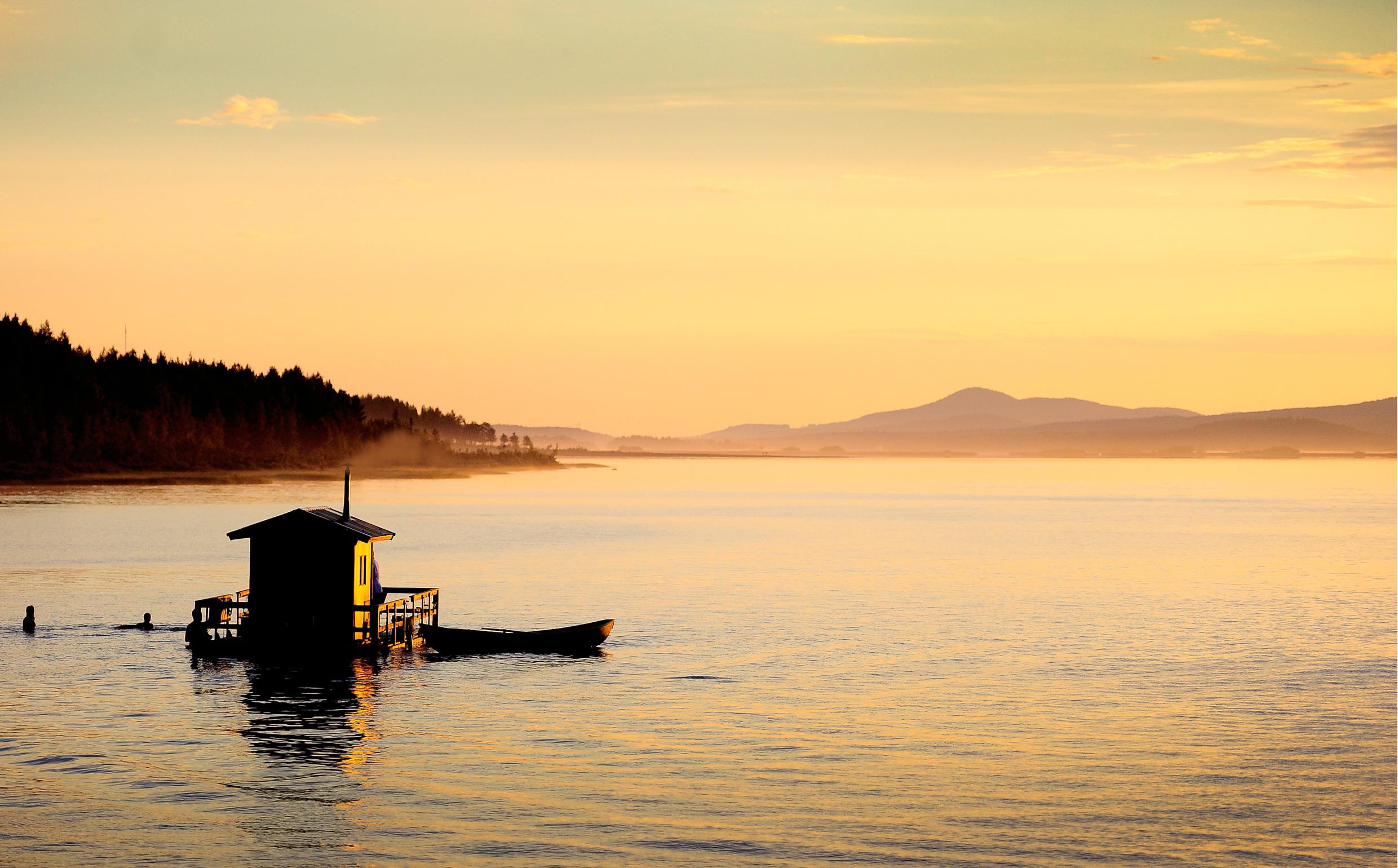 Un bateau-sauna sur un lac au coucher du soleil. Les gens se baignent près du sauna. Une barque est amarrée au bateau-sauna.