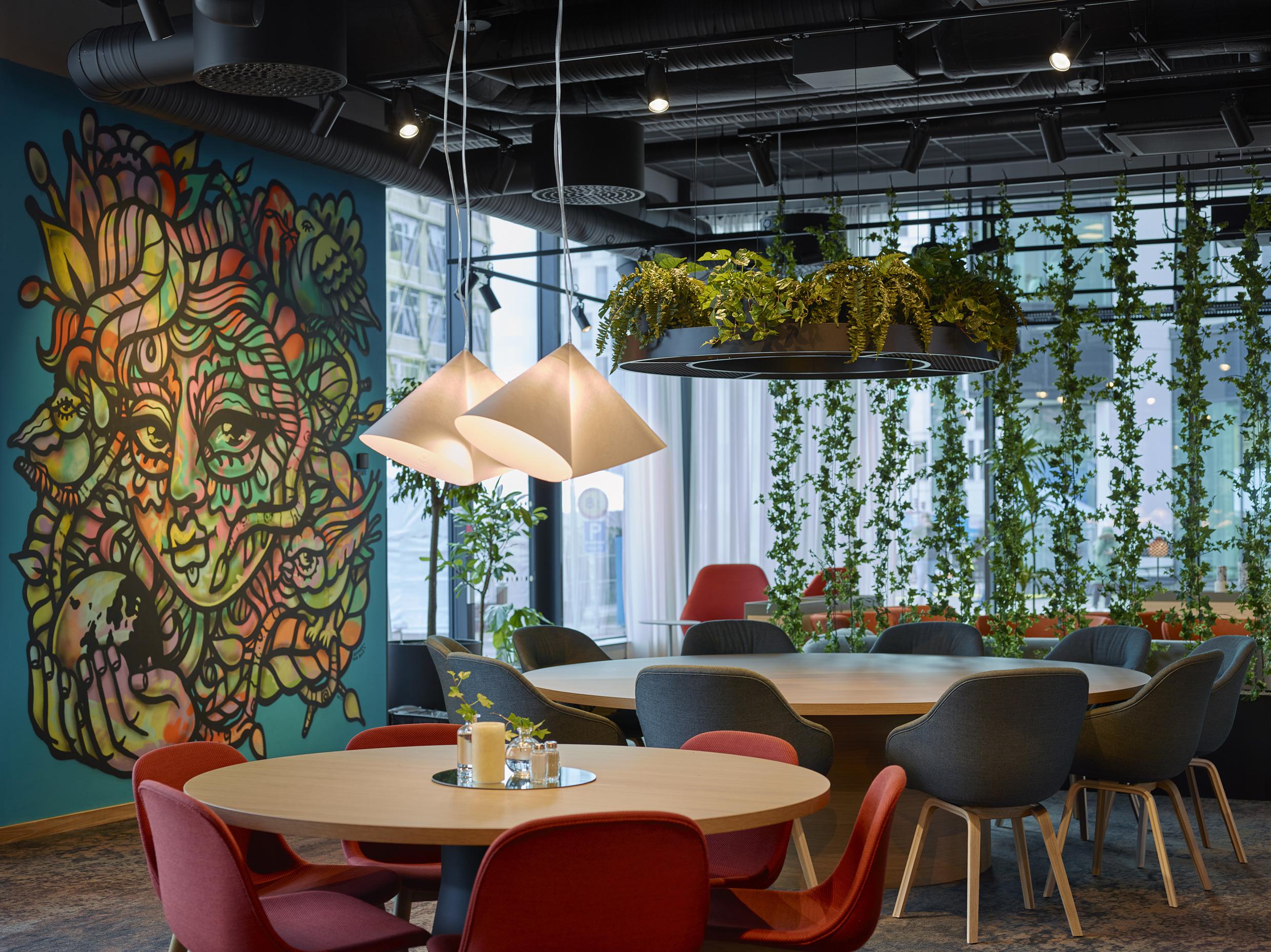 Een kamer met ronde houten tafels met rode en zwarte stoelen. Aan het plafond hangen planten en een blauwe muur is beschilderd met kunstwerken.