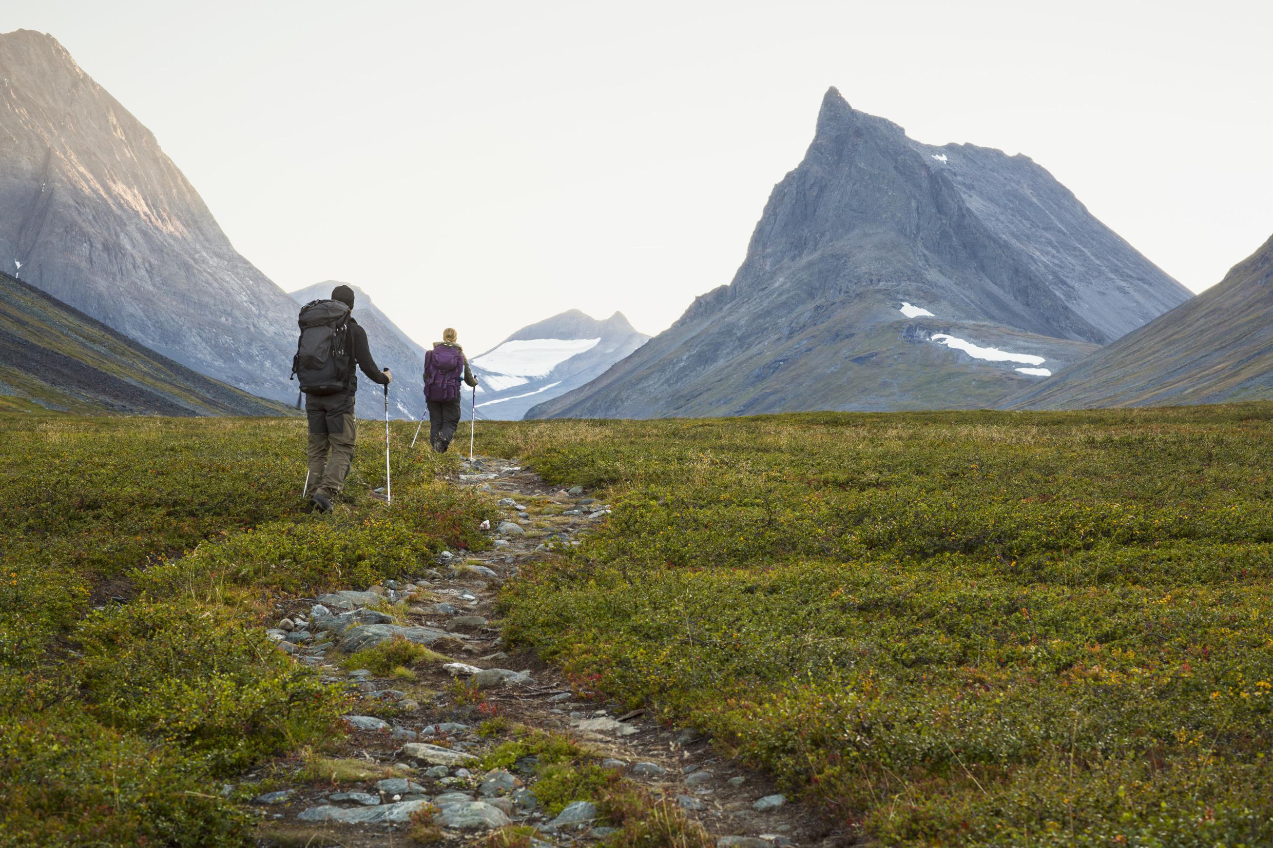 Zwei Wanderer wandern mit ihren Wanderstöckern auf einen begrasten Weg, der durch karge, steile Berge führt.