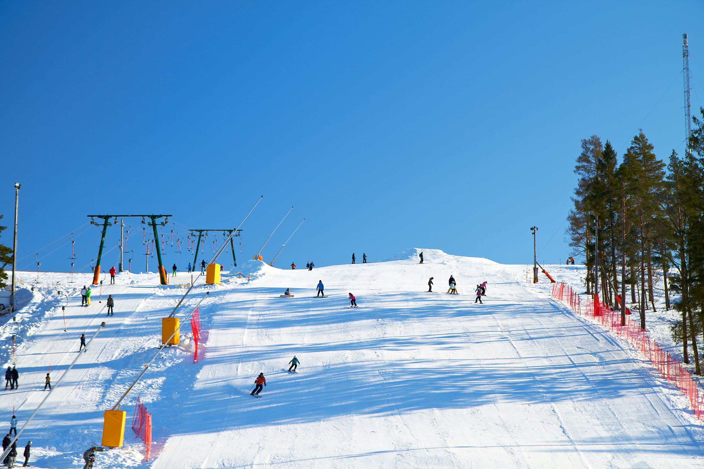 Skiers on a sunlit slope at Alebacken, with button lifts and pine trees under a clear blue sky.