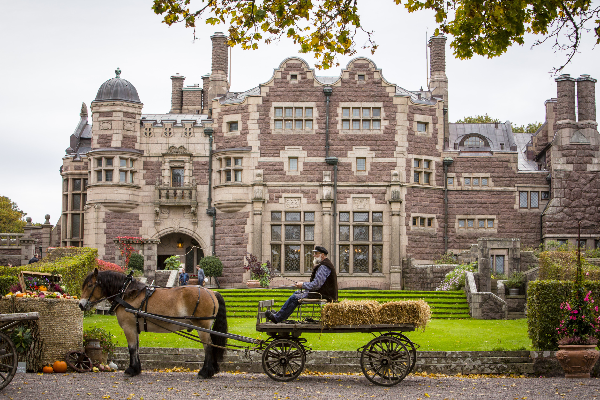 An elderly man is driving a horse and carriage with a couple of hay bales. They stand in front of a castle and beside you see a market stall with vegetables.