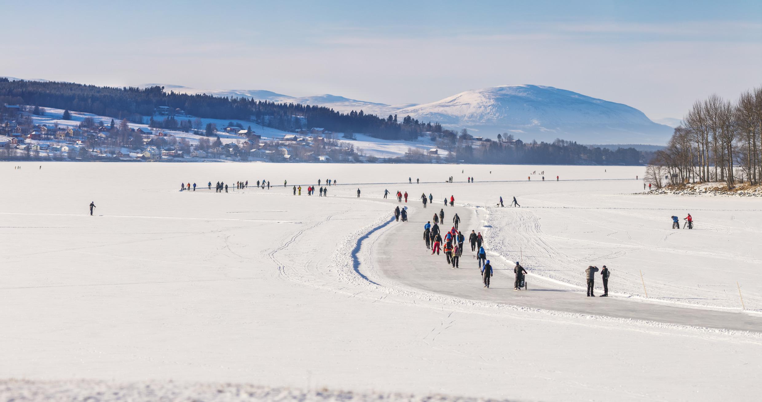 Verschillende mensen schaatsen op geprepareerde banen op een met sneeuw bedekt bevroren meer. Op de achtergrond staan huizen, bomen en bergen.