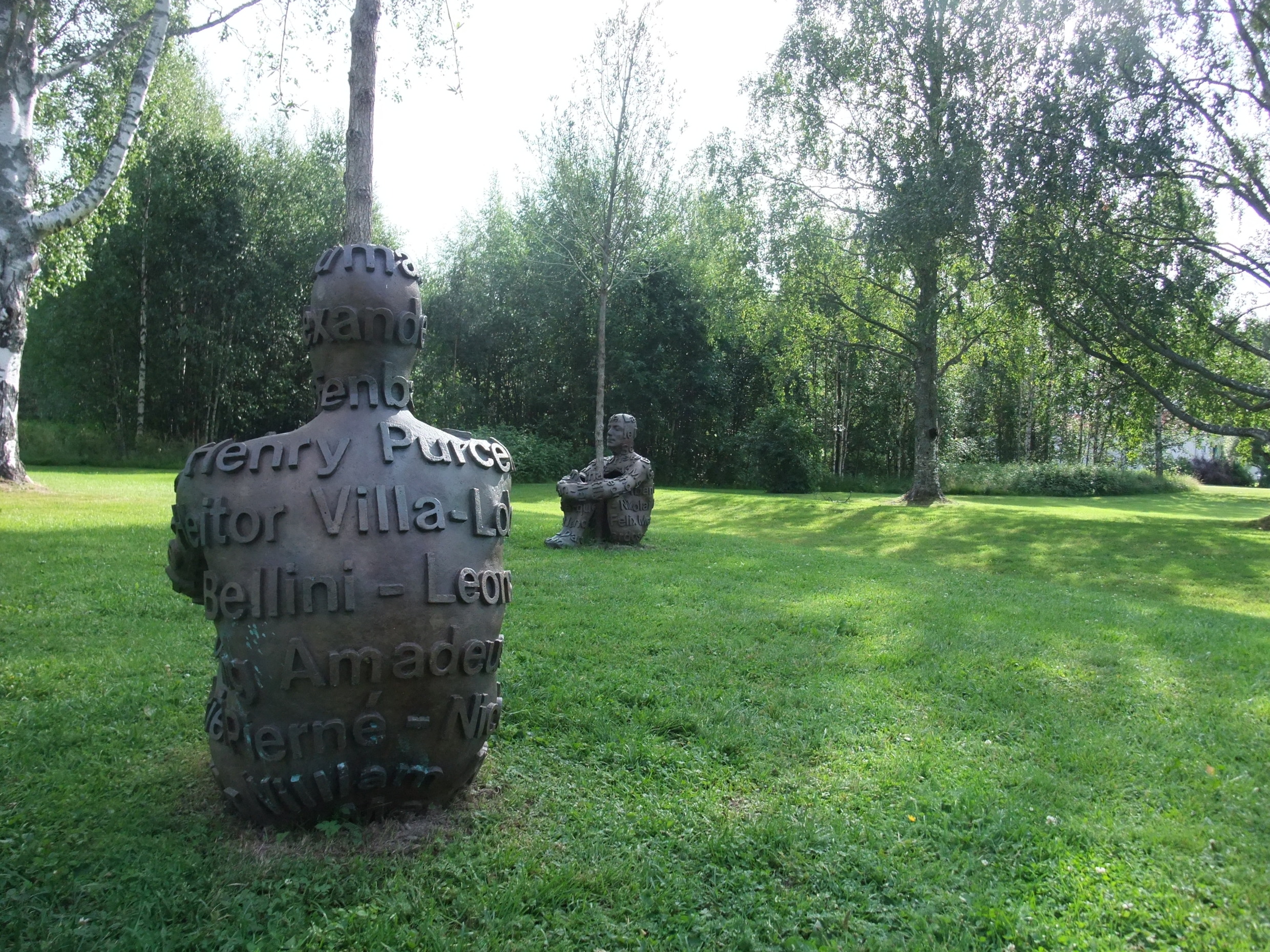 Outdoor sculptures of seated human figures in a grassy, tree-filled park at Umedalen Sculpture Park in Umeå, northern Sweden.