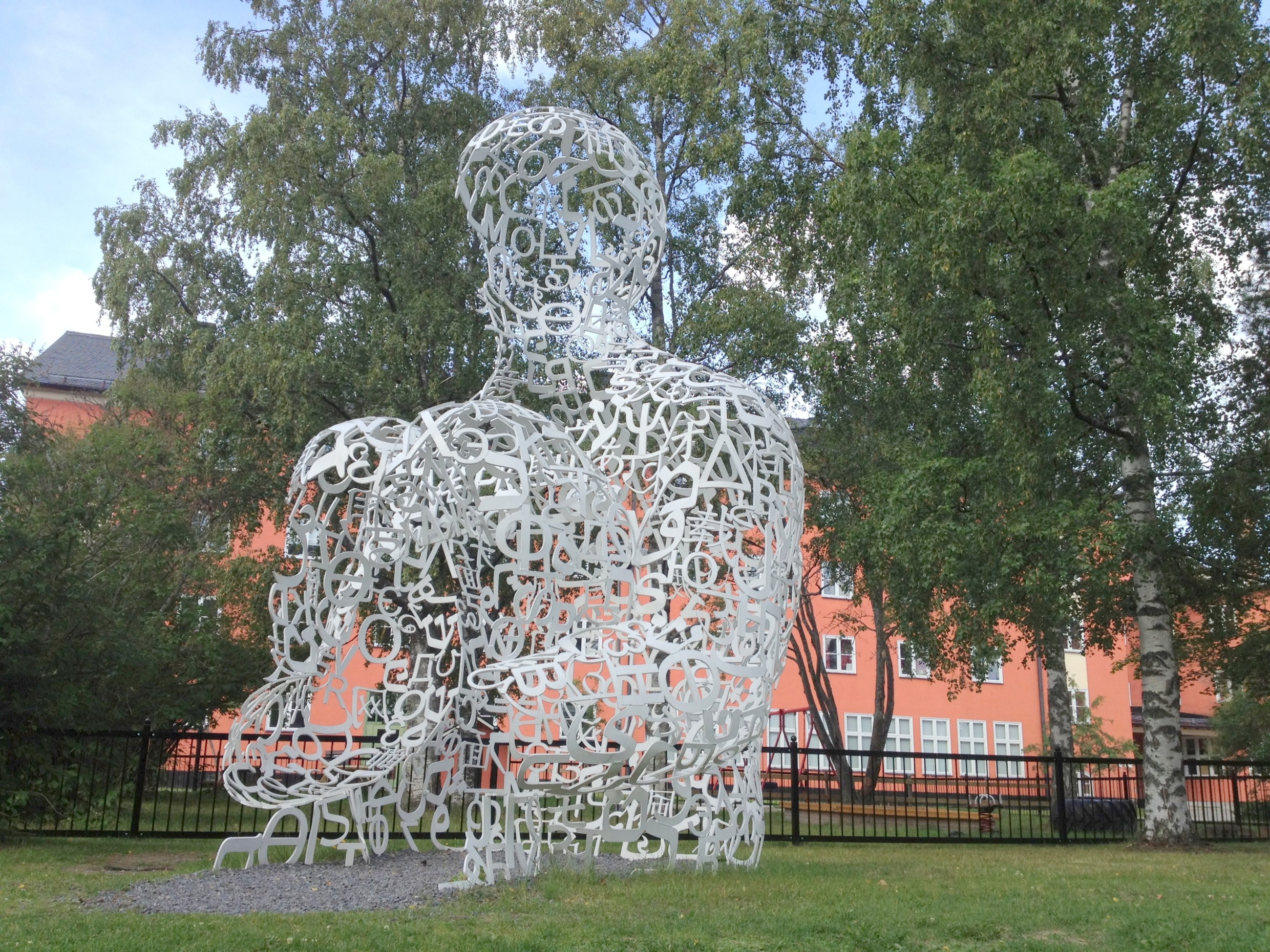 Une sculpture composée de lettres blanches réalisée par Jaume Plensa dans le parc de sculptures d'Umedalen, à Umeå, avec des arbres et un bâtiment historique orange en arrière-plan.