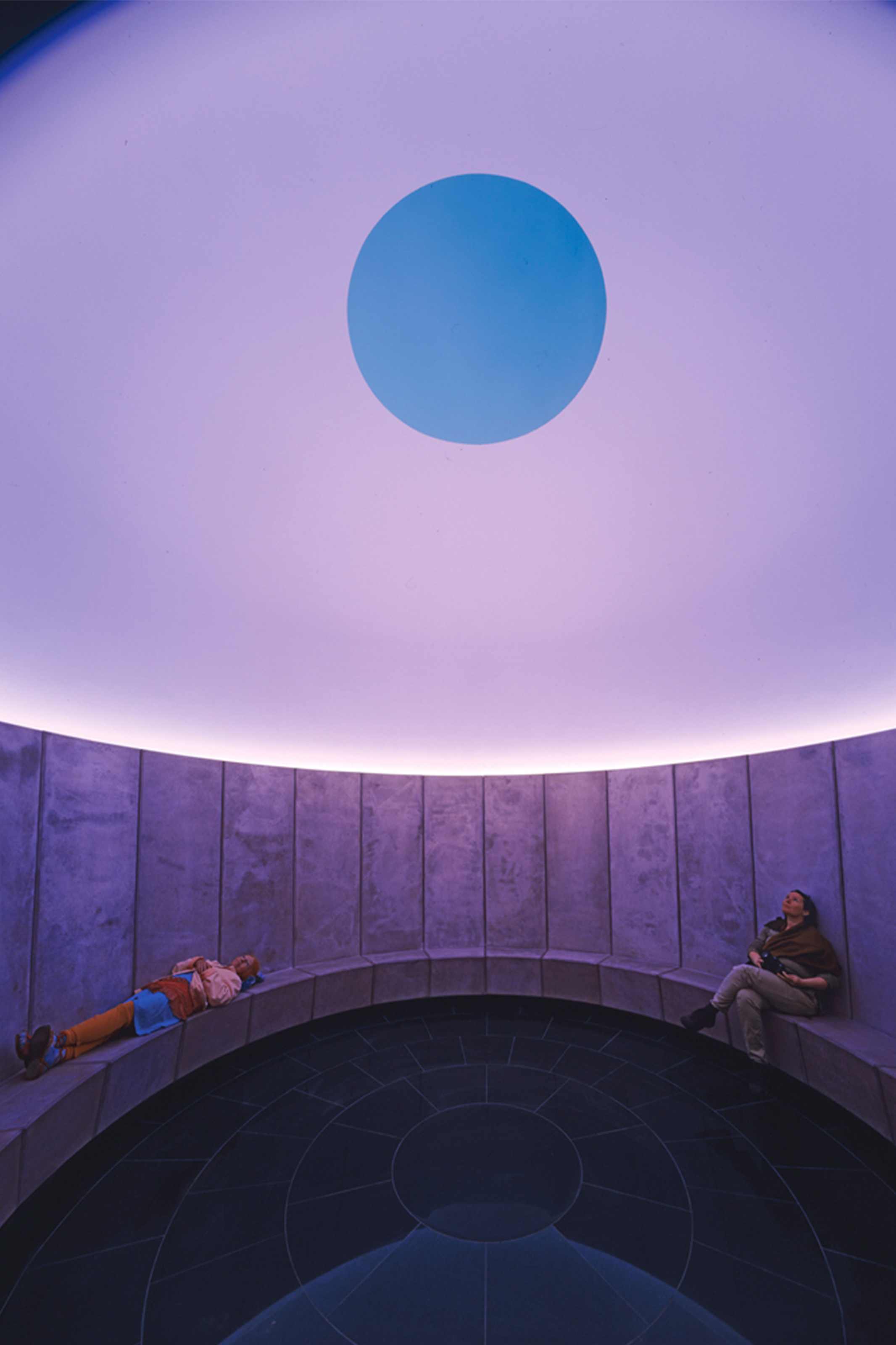 Deux personnes assises à l'intérieur du Skyspace de James Turrell à Ytterjärna, en Suède, sous un dôme lumineux avec une ouverture circulaire vers le ciel.