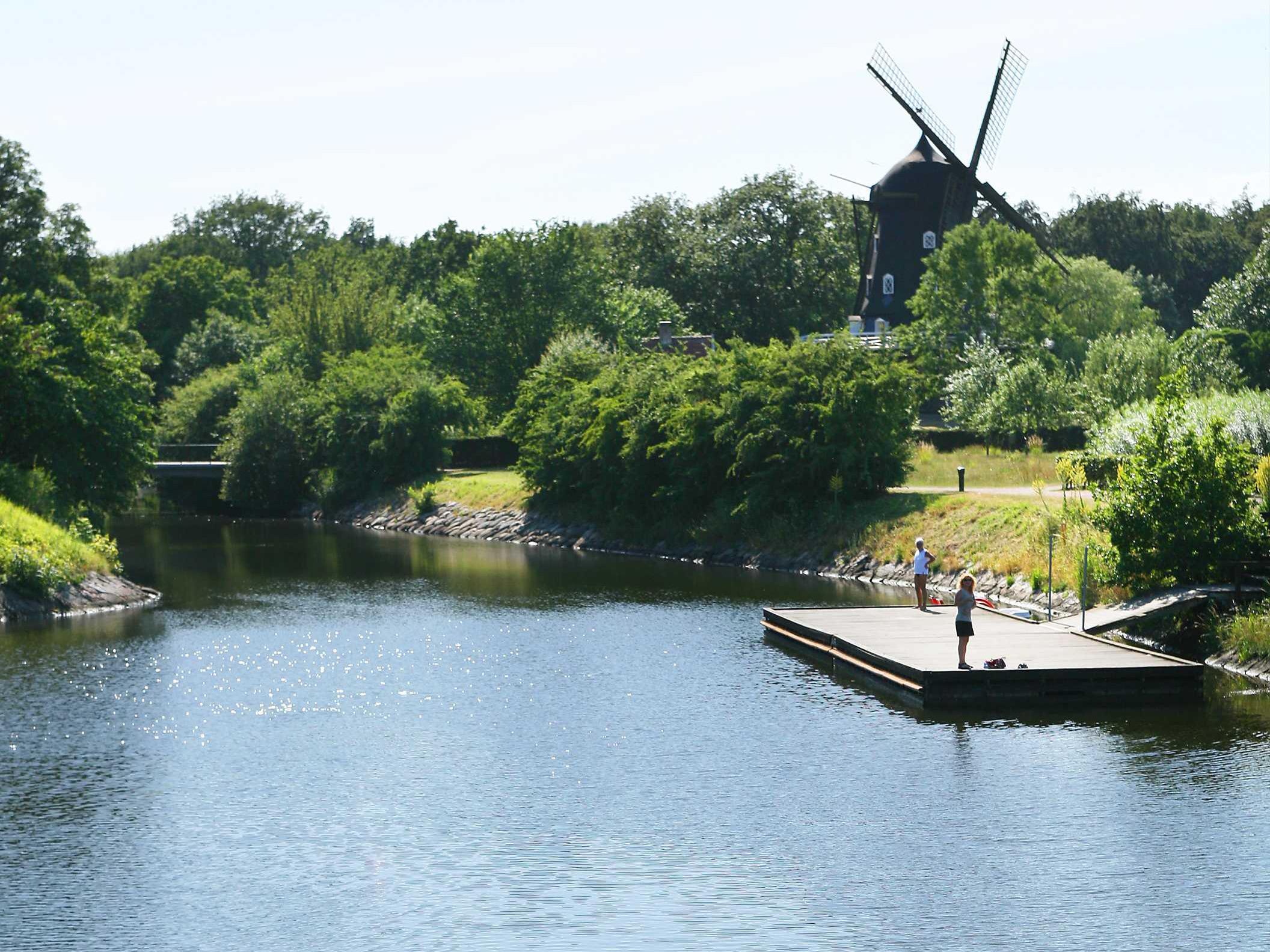 Twee vrouwen staan op een steiger aan een gracht in een stadstuin. Op de achtergrond staat een windmolen omgeven door groen.