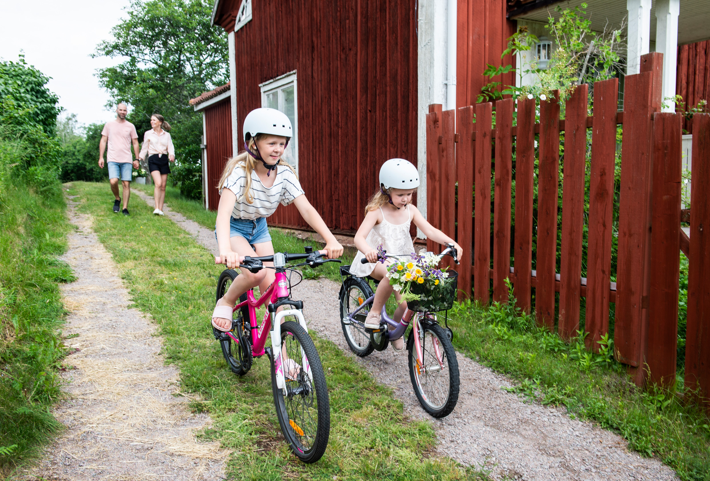 Zwei Kinder fahren mit dem Fahrrad auf einer kleinen Straße neben einer roten Hütte. Ein Mann und eine Frau gehen hinter ihnen her.