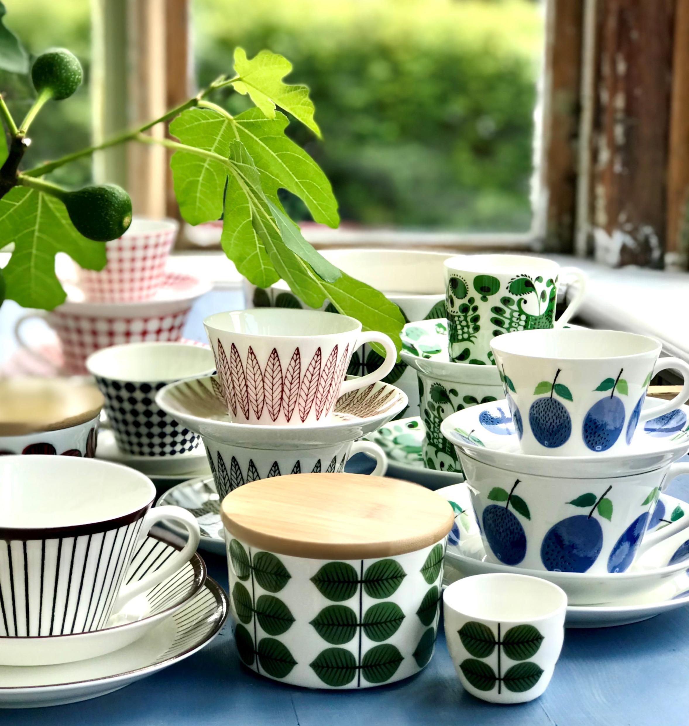 Close-up on porcelain coffee cups with different patterns on a table and a twig from a green plant.