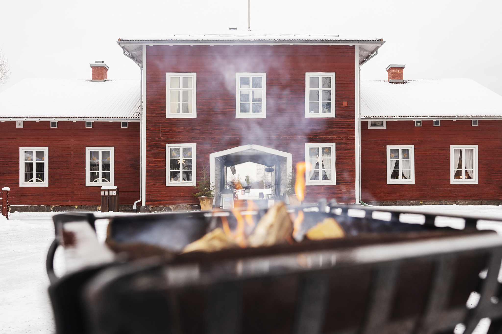 Red wooden building at Stenegård in winter, with snow-covered ground and fire burning in the foreground.