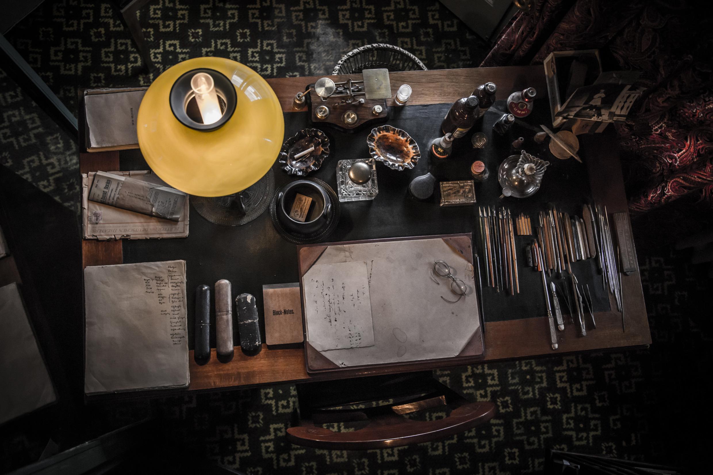 An old wooden desk, with pencils lined up, paper and notes, glasses and an old lamp.