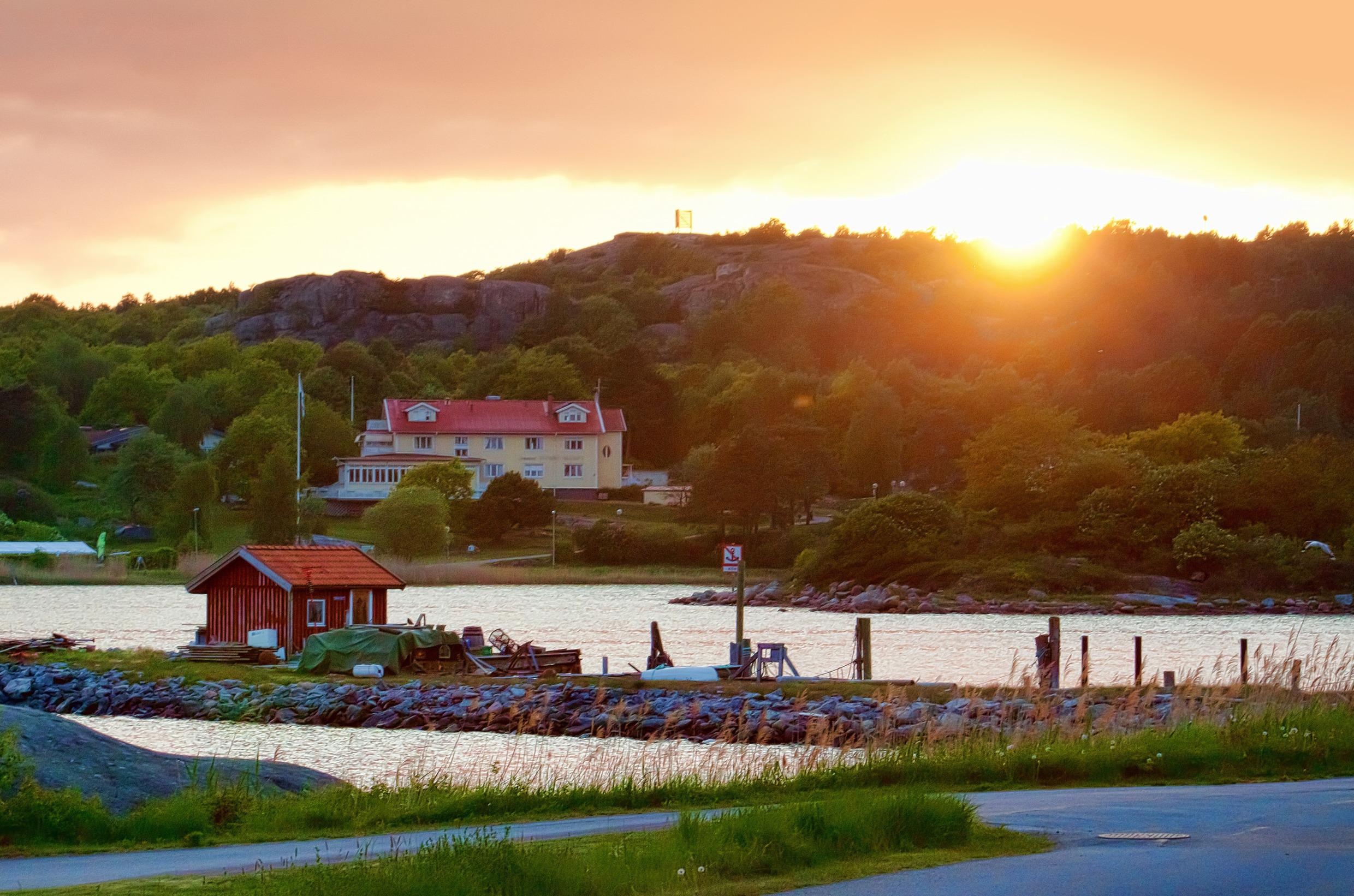 Een heuvelachtige zeekust begroeid met groen. Op de achtergrond een huis en een ondergaande zon.