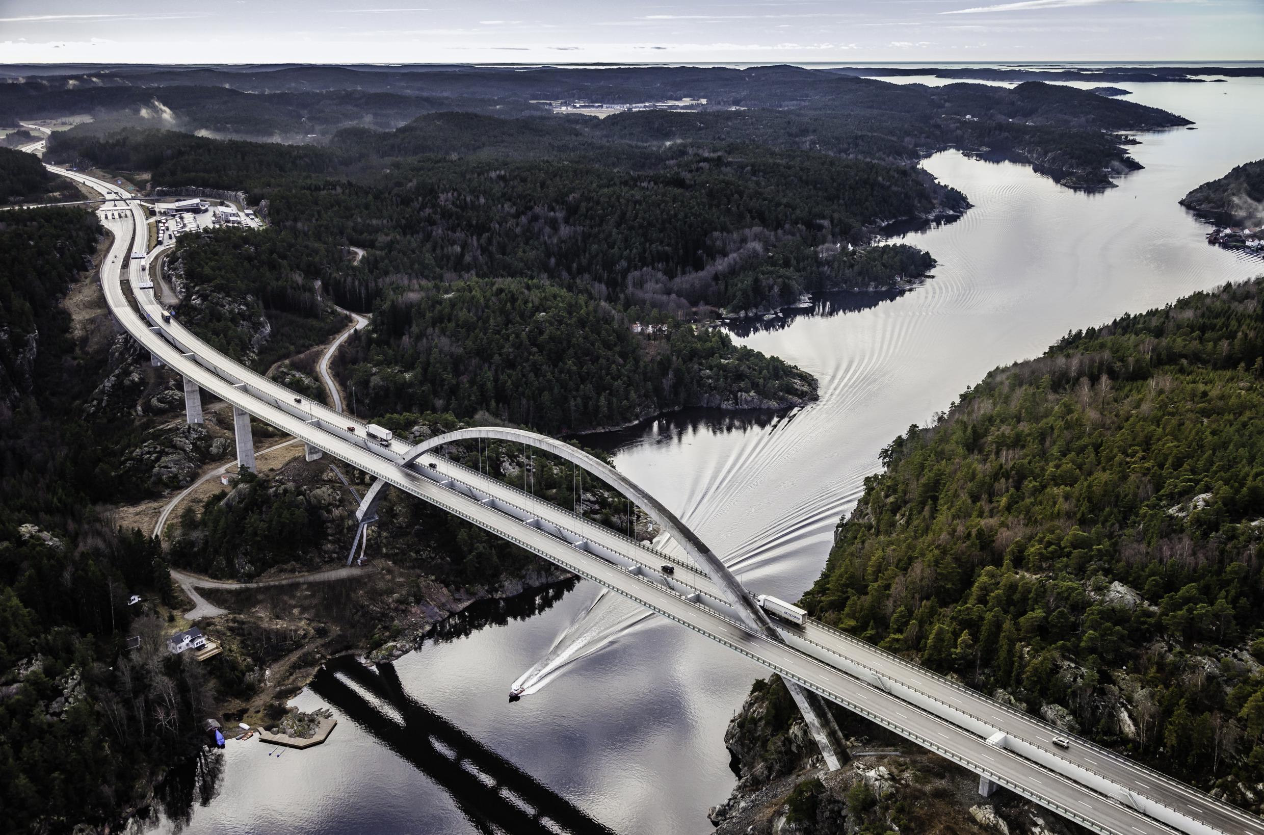 Auf der Svinesund-Brücke herrscht reger Verkehr, und unter der Brücke rast ein Motorboot. Die Landschaft auf beiden Seiten der Brücke ist hügelig.