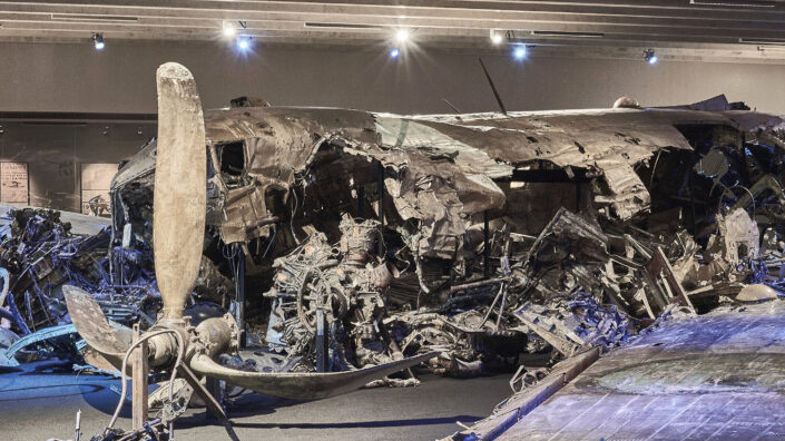 De verfrommelde resten van een DC-3-vliegtuig uit de tijd van de Koude Oorlog, tentoongesteld in het Zweedse Luchtmachtmuseum in Linköping, met gedempte verlichting en dramatische schaduwen.