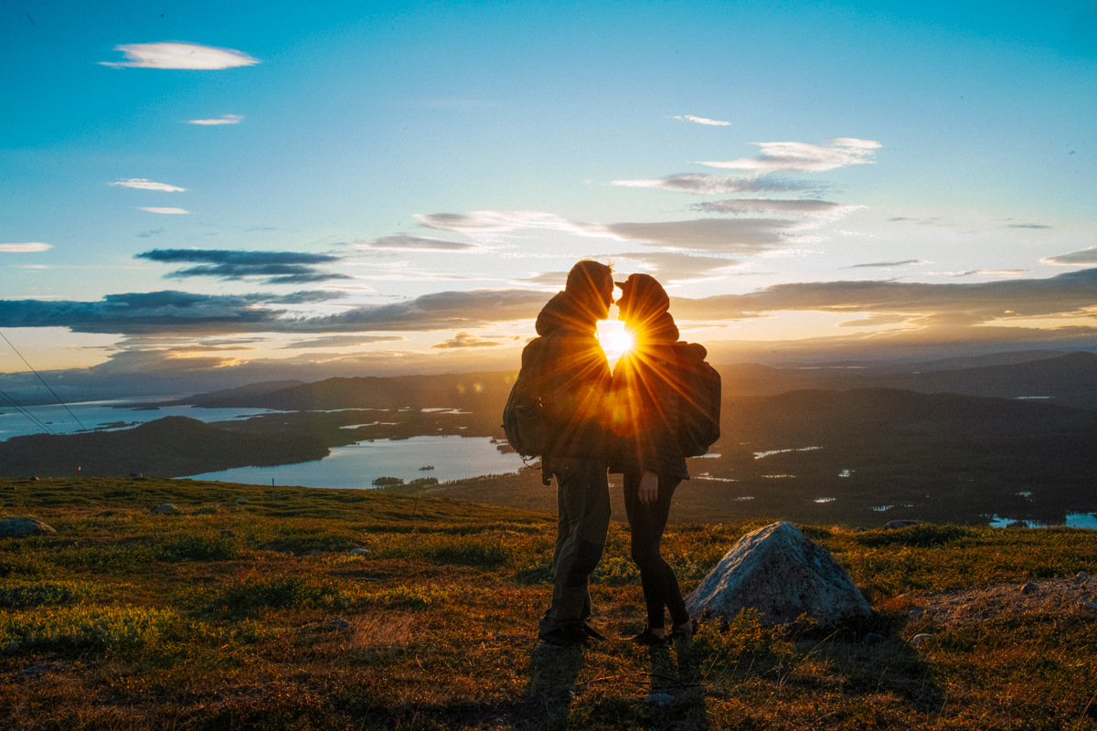 Two hikers standing on a mountain with the sun low on the horizon, overlooking lakes and landscapes in Arctic Sweden during the Midnight Sun.