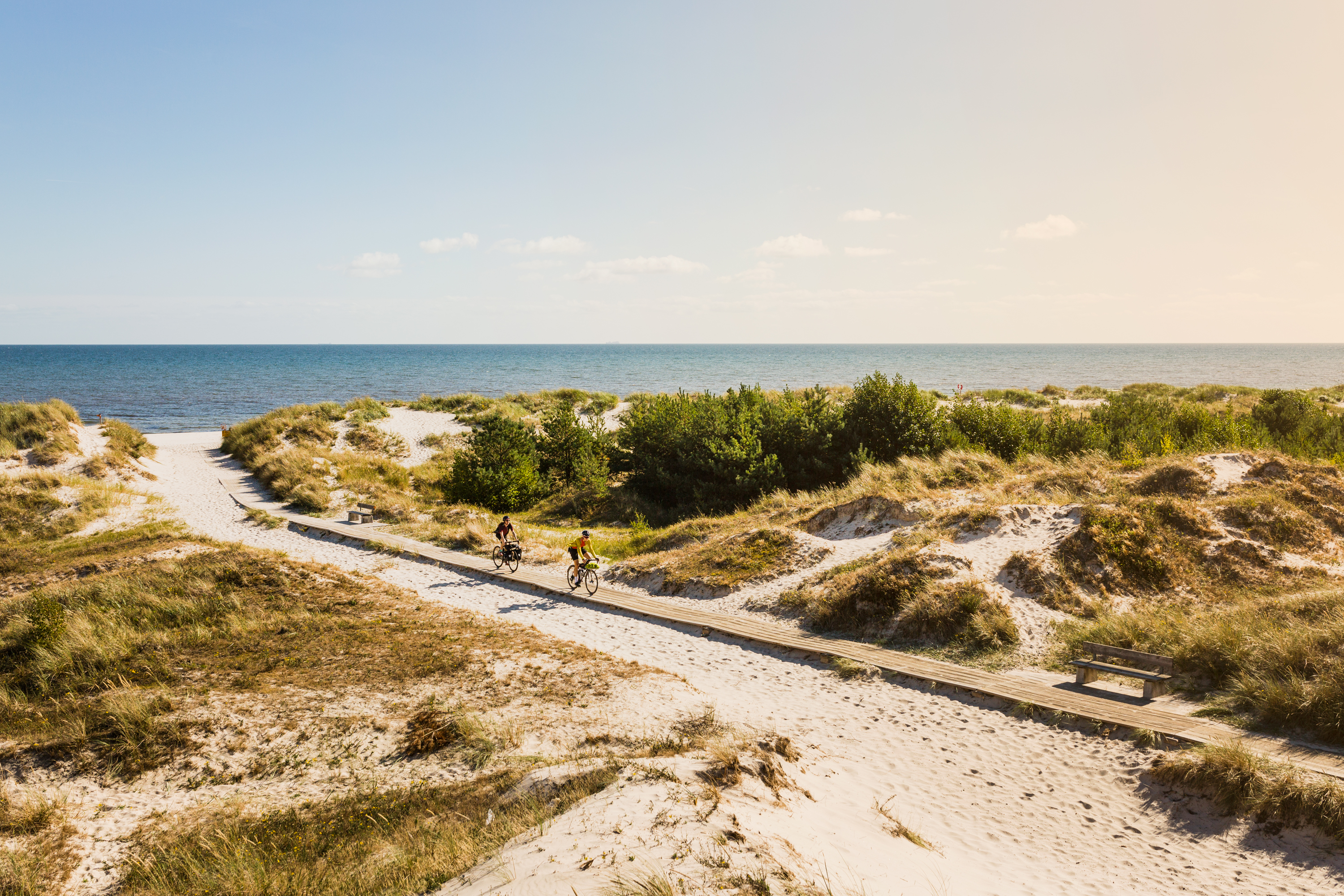 Twee mensen die fietsen op een houten loopbrug op een zandstrand met hier en daar wat gras.