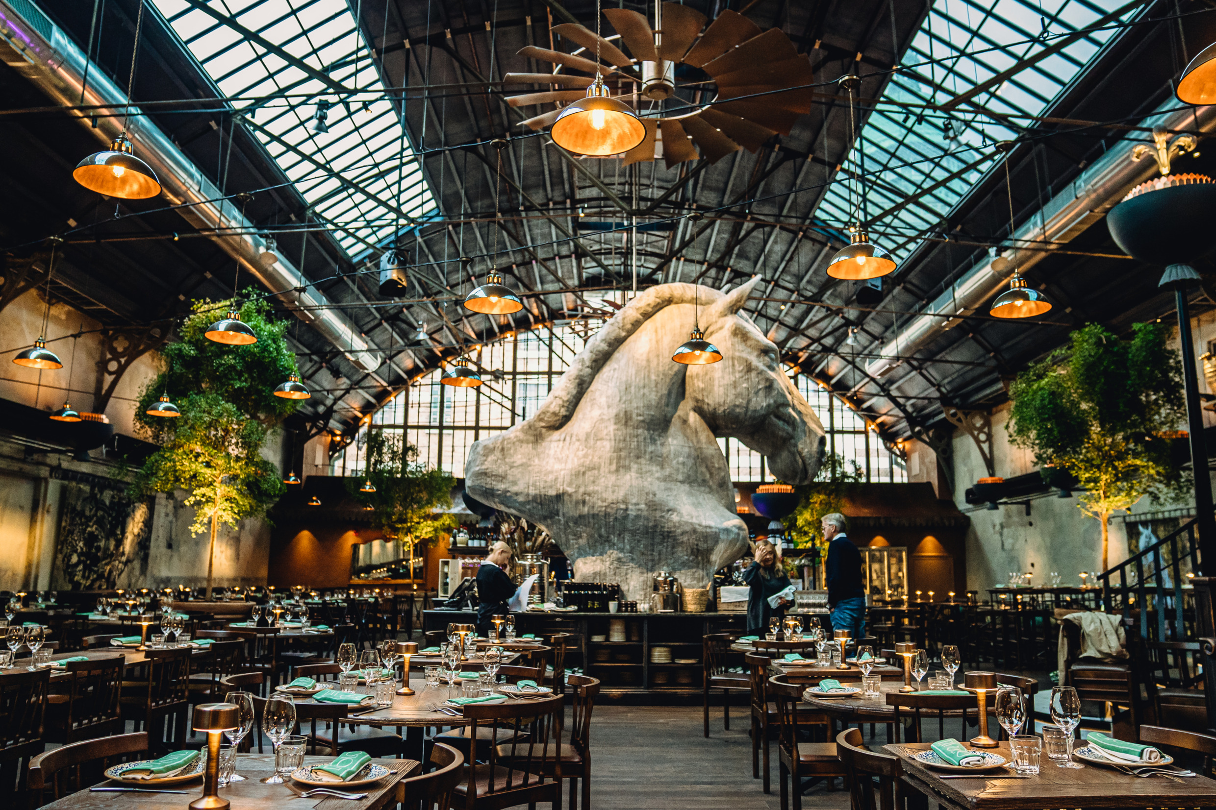 Intérieur du restaurant Tavolo à Göteborg, avec une sculpture de cheval imposante, un éclairage de style industriel et des tables en bois élégamment disposées dans une salle à manger spacieuse.