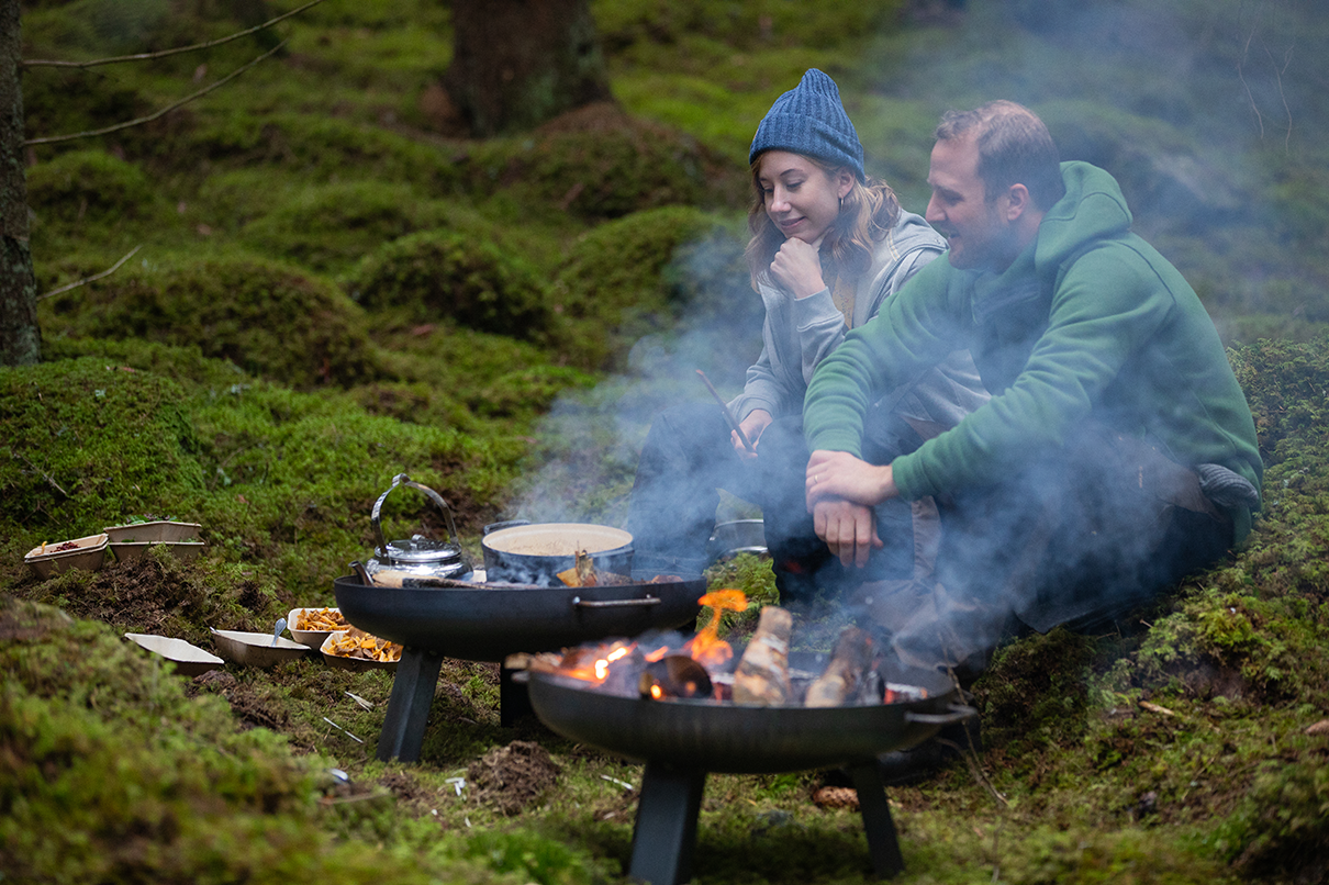 Un homme et une femme assis dans la forêt sont en train de cuisiner sur des feux ouverts. De la fumée s'échappe.