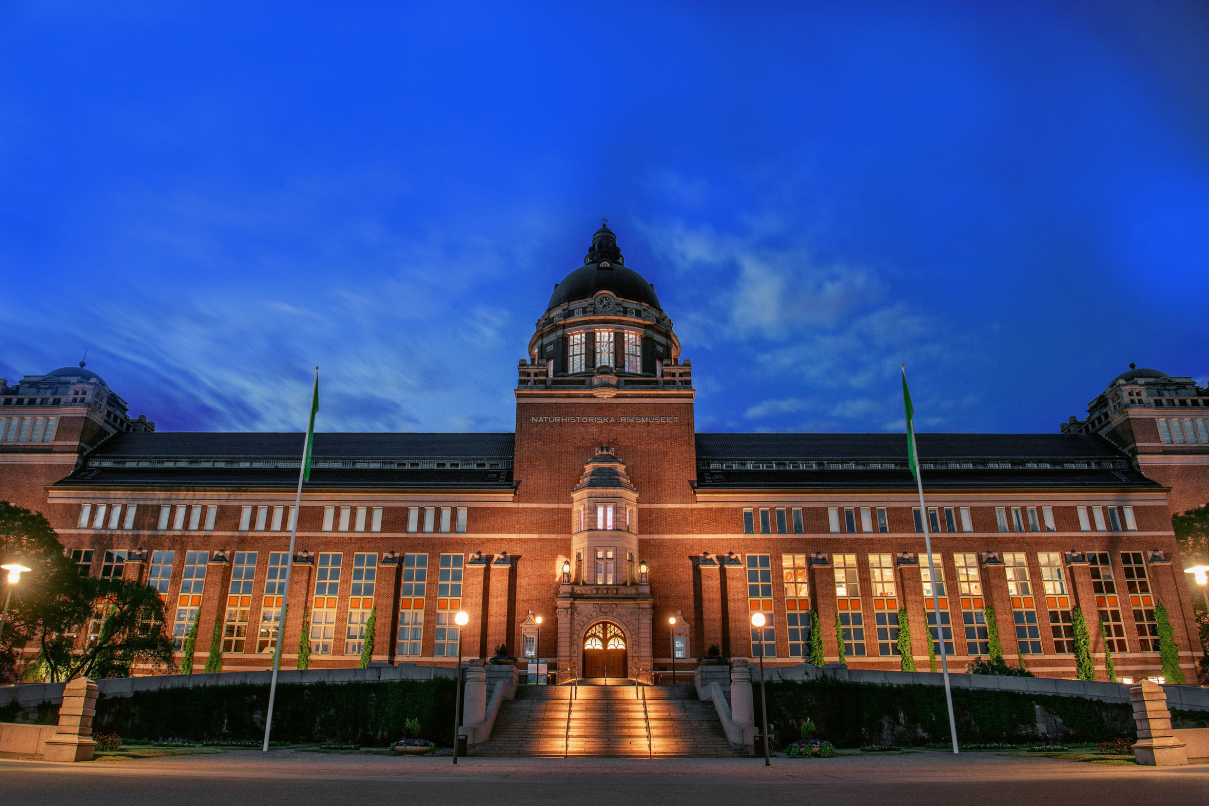 Nachtfoto van de verlichte gevel van het Natuurhistorisch Museum in Stockholm, met een koepeldak en symmetrische architectuur onder een schemerige hemel.