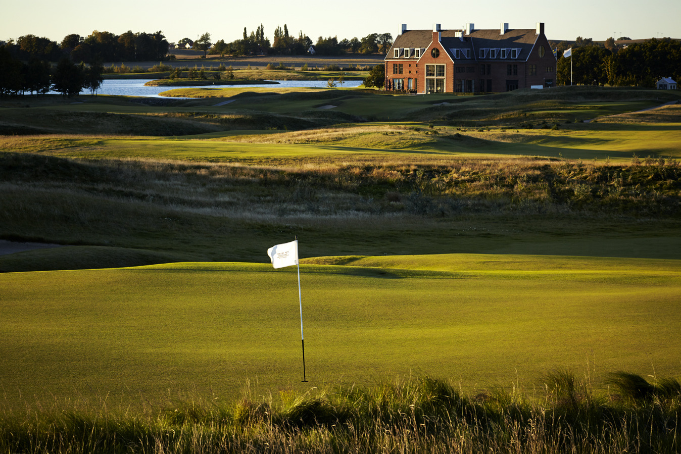 Golf course at sunset with a house in the background and a putting green with a flag in the foreground.