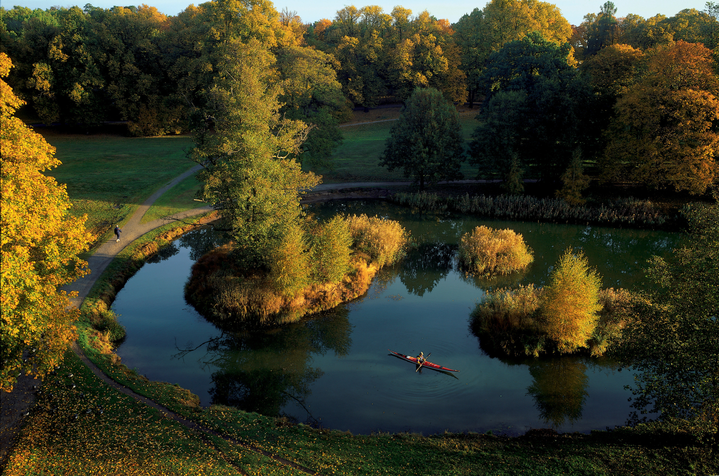 Person paddling a red kayak on a small lake surrounded by autumn trees in Haga Park, Stockholm.