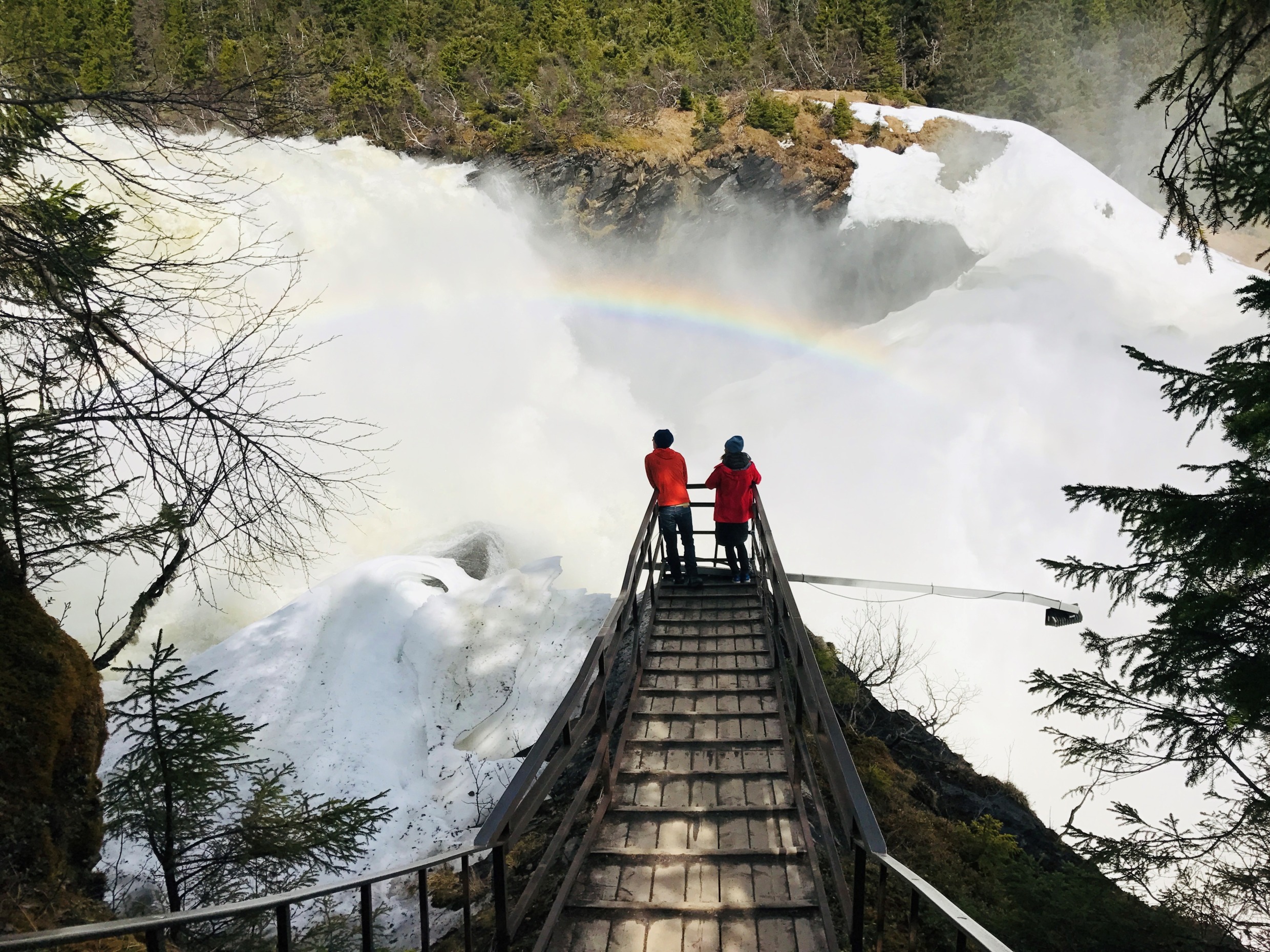 Two people overlook a huge waterfall. The water from the waterfall creates a rainbow.