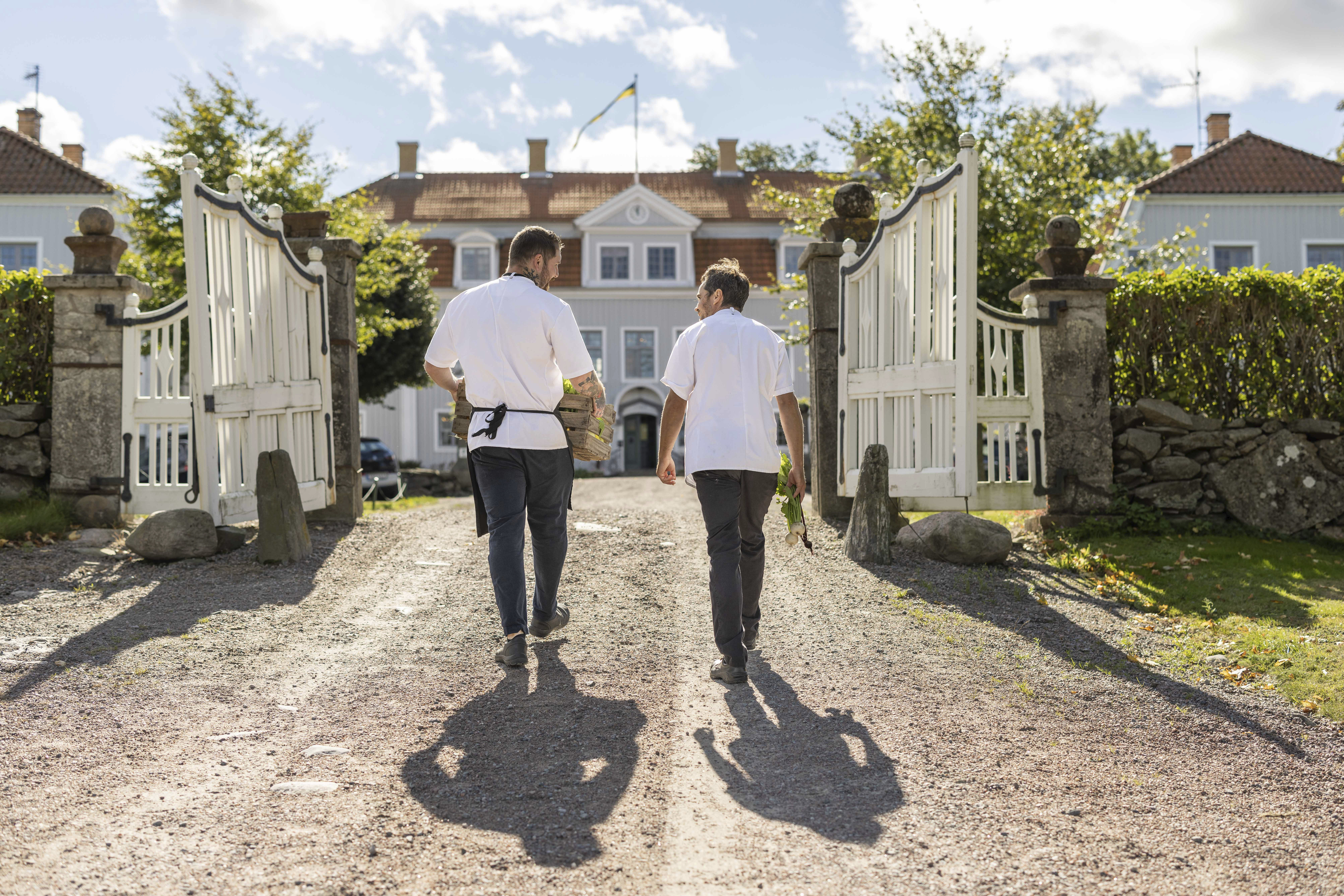 Deux membres du personnel franchissent l'entrée du manoir de Tofta, chargés de produits frais, avec le manoir en arrière-plan.