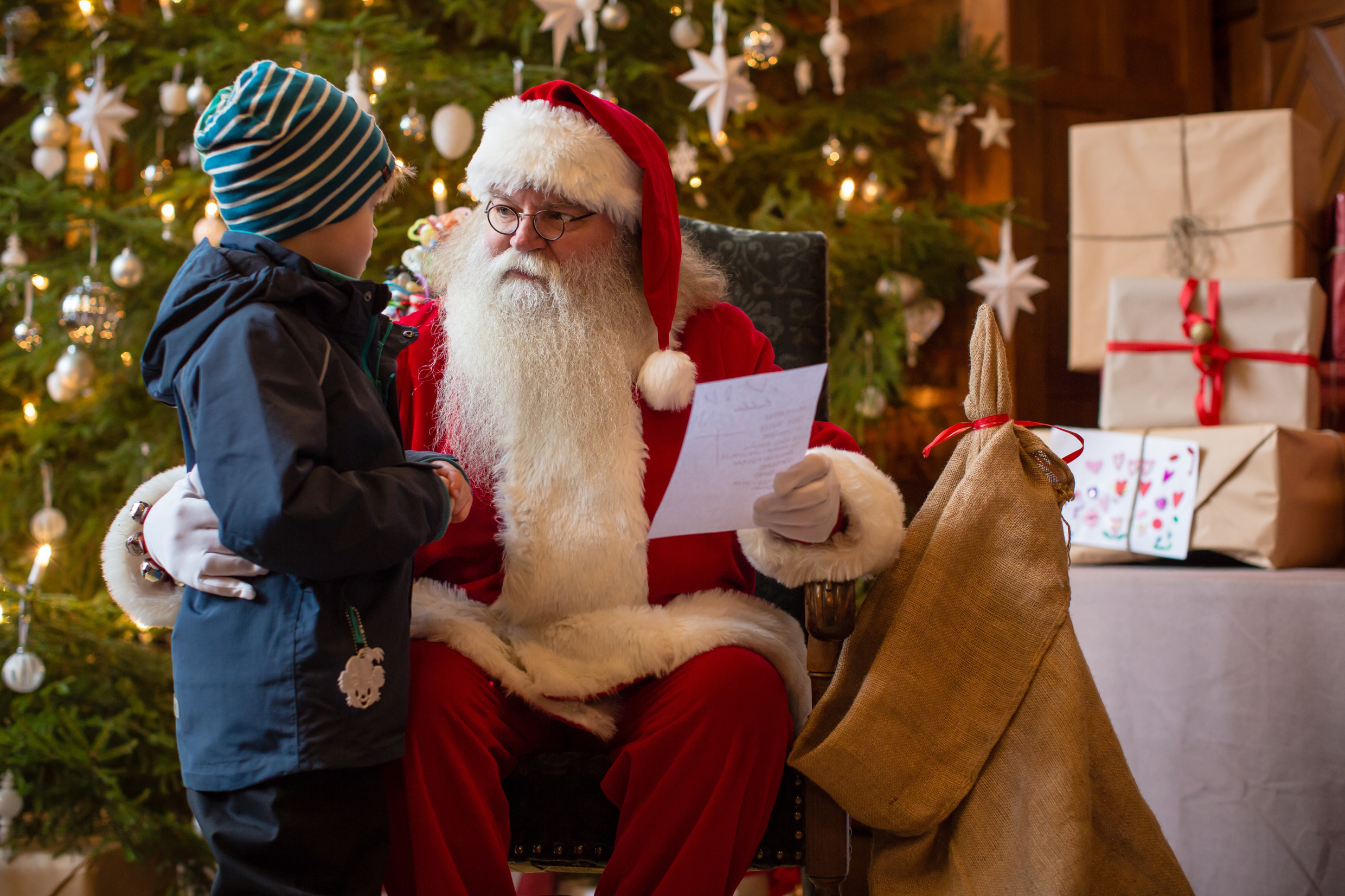 De kerstman, een oude man met een witte baard en een rode mantel, zit voor een kerstboom en praat met een kind.