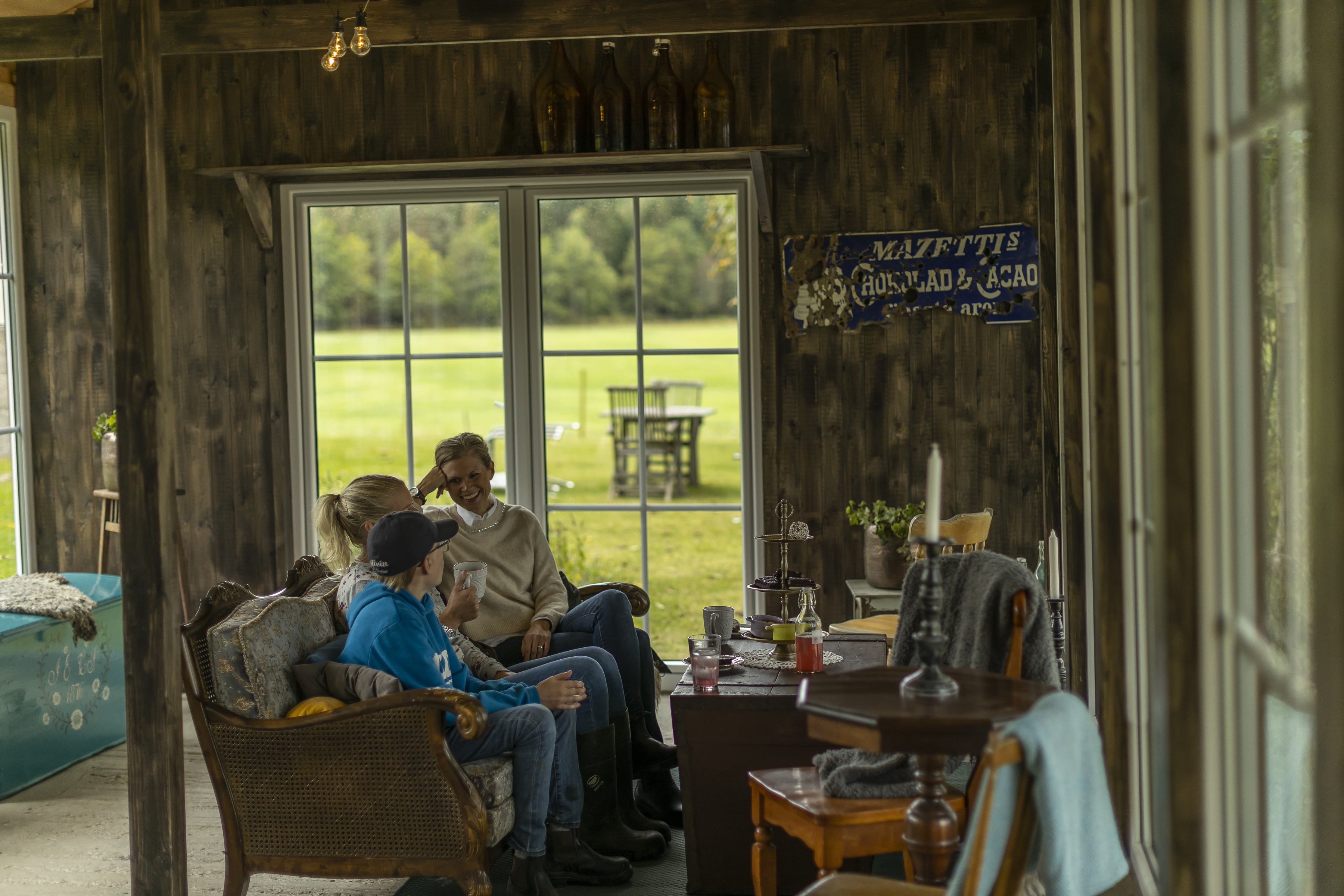 Three people sitting and chatting in a rustic indoor setting at Farmlodge, with large windows overlooking a green field outside.