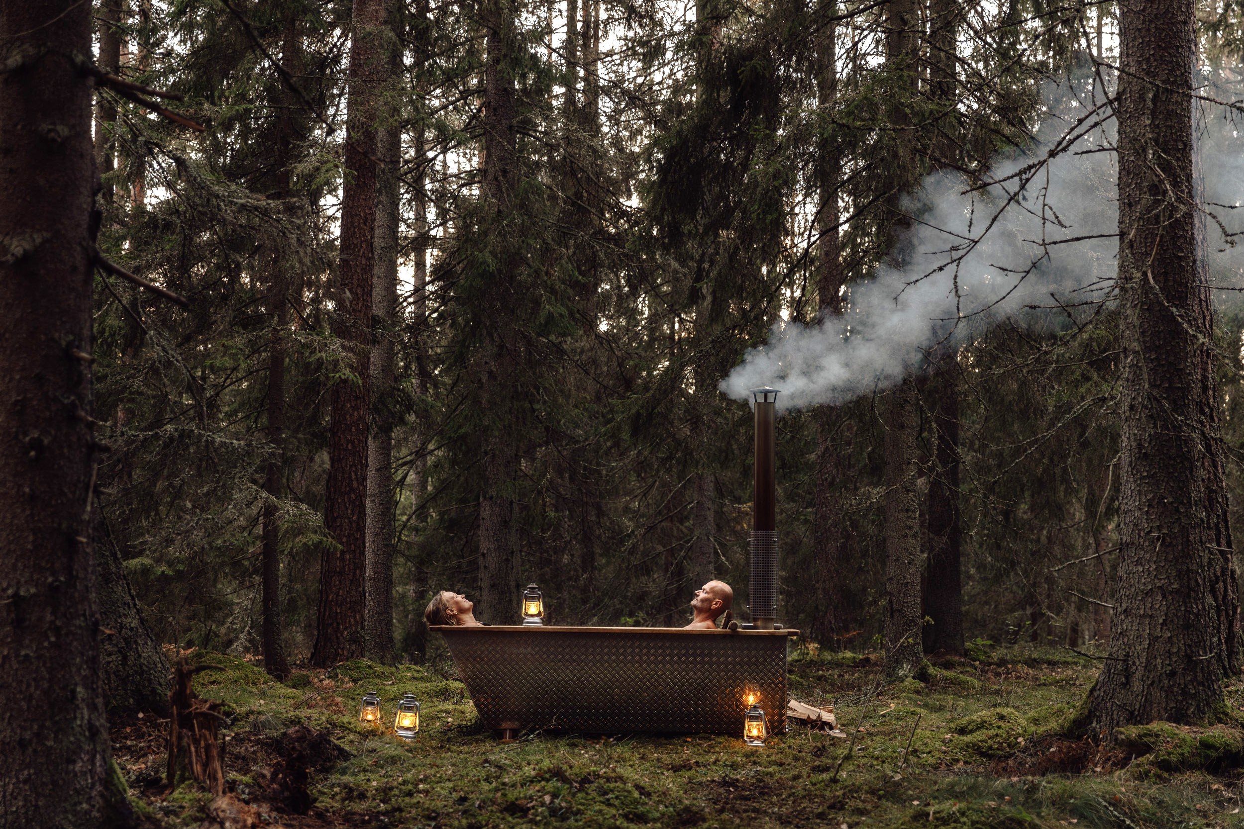Two people relaxing in a wood-fired outdoor bathtub in a forest, with lanterns placed around them and smoke rising from the stove.