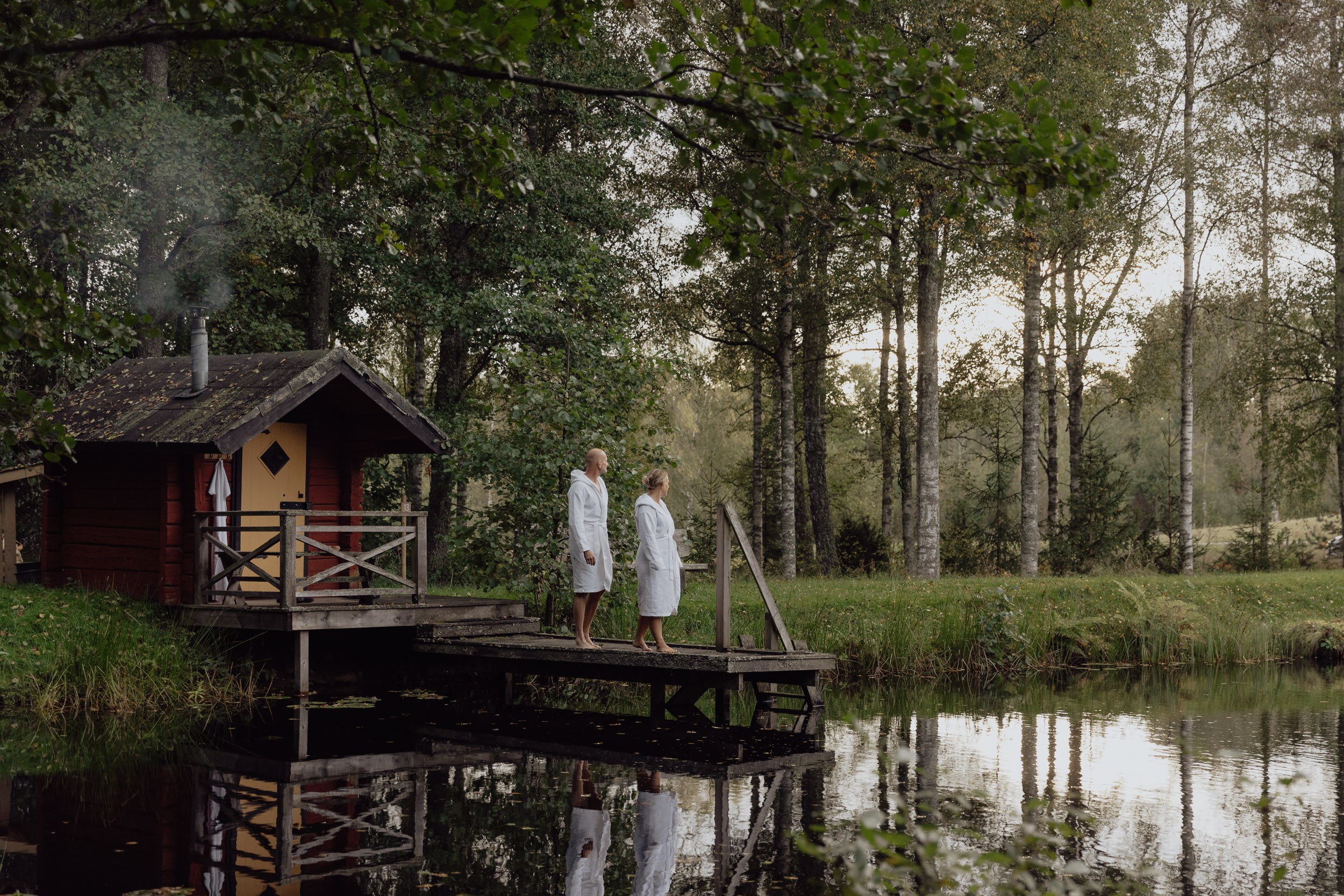 Two people in white robes standing on a wooden jetty beside a small lakeside sauna in a forest setting.