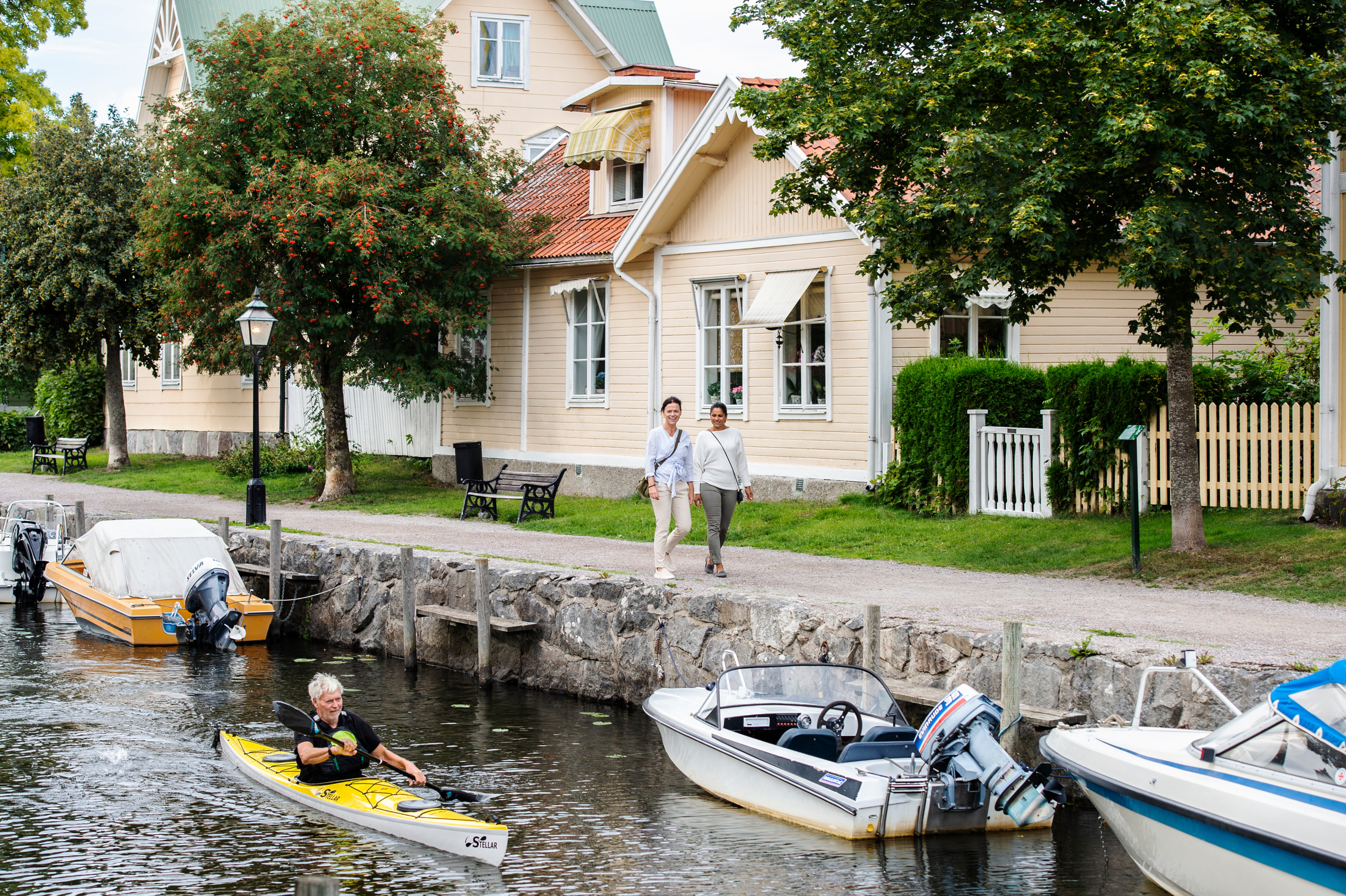 Un homme fait du kayak sur un canal dans une petite ville, où se trouvent de petits bateaux. Deux femmes marchent le long du canal.