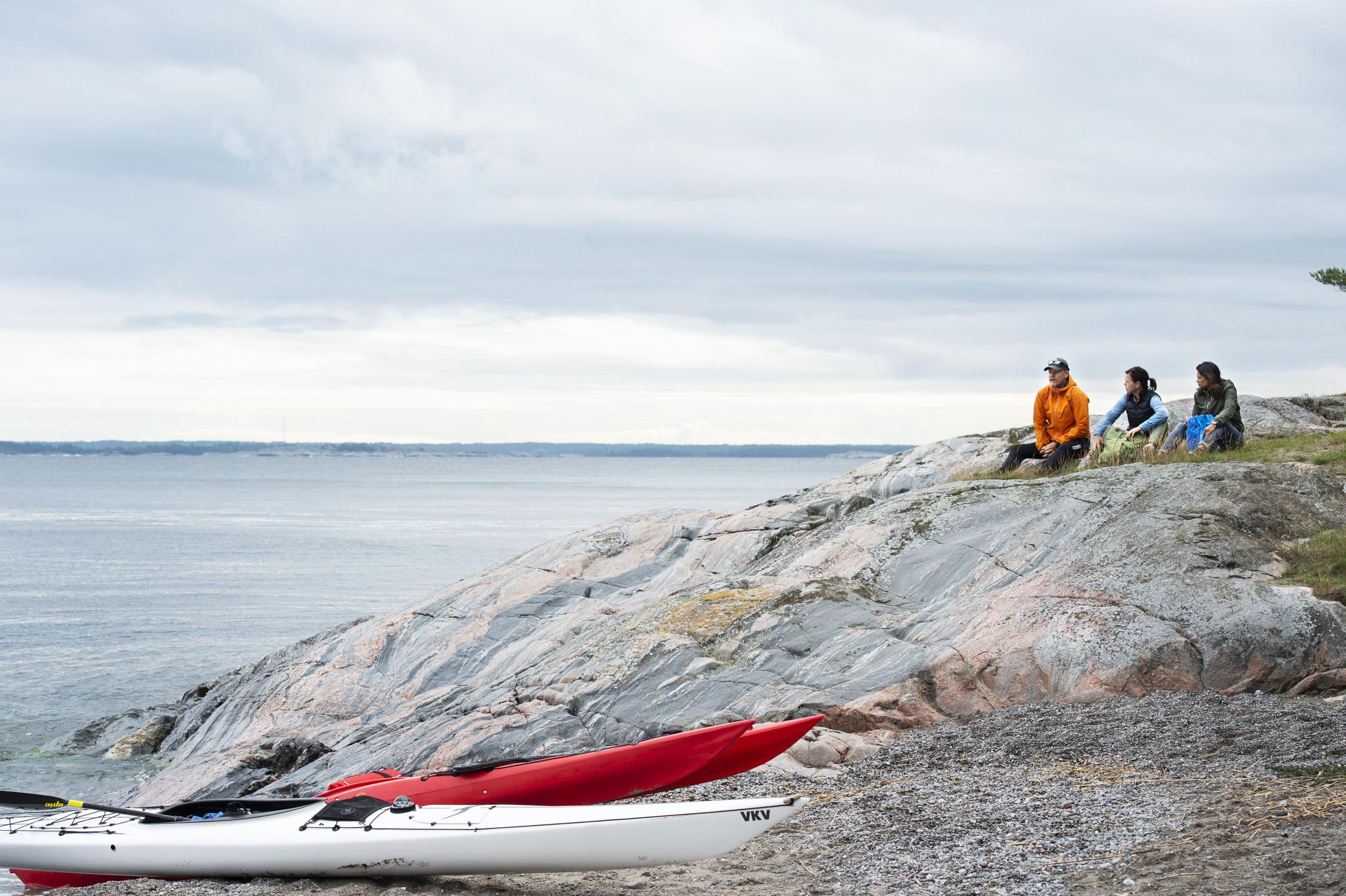 Ein Mann und zwei Frauen sitzen auf einer Insel im Archipel. Drei Kajaks liegen am Wasser.