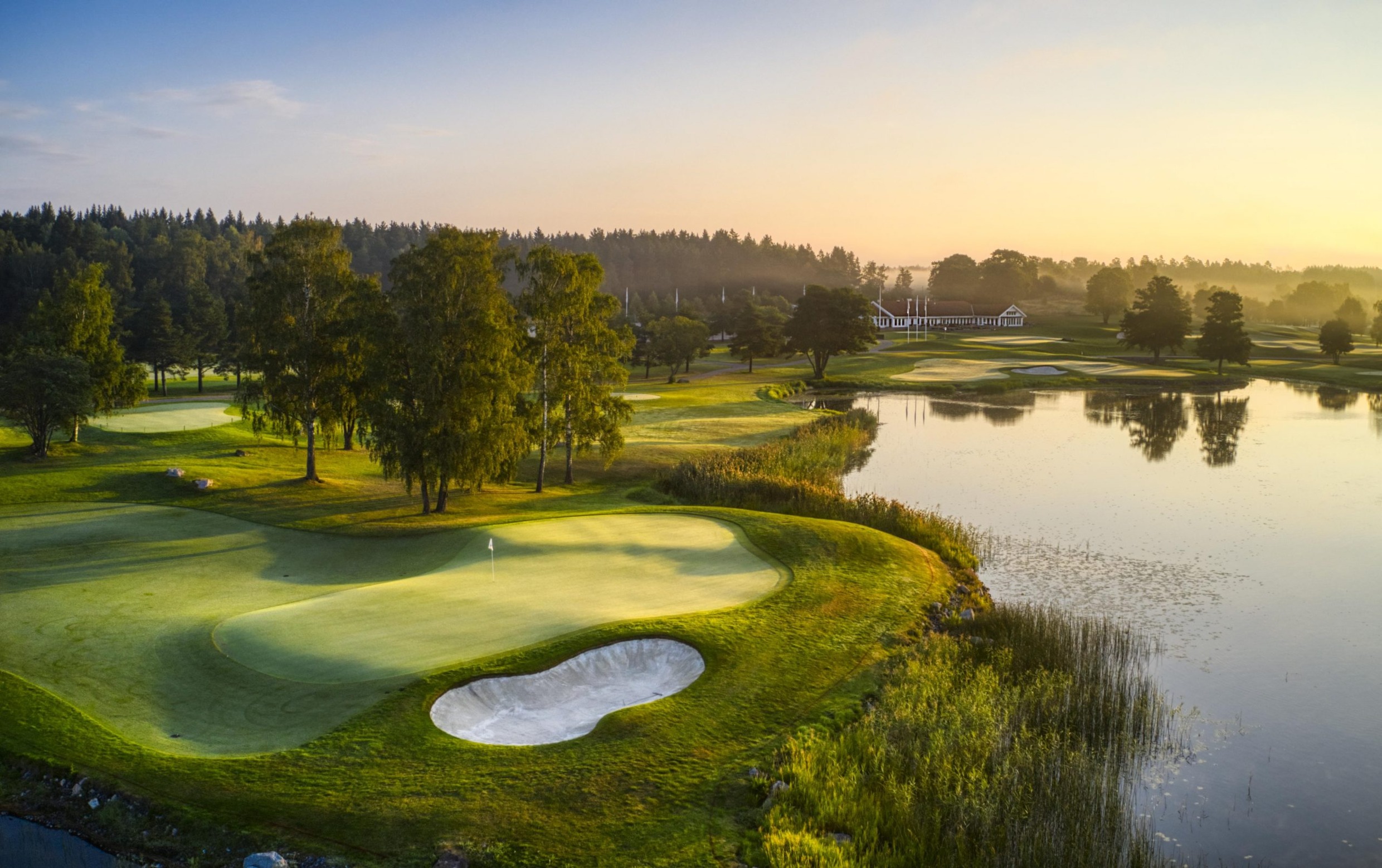 View over Ullna Golf Course early in the morning, with bunkers, a lake, and green fairways surrounded by trees.