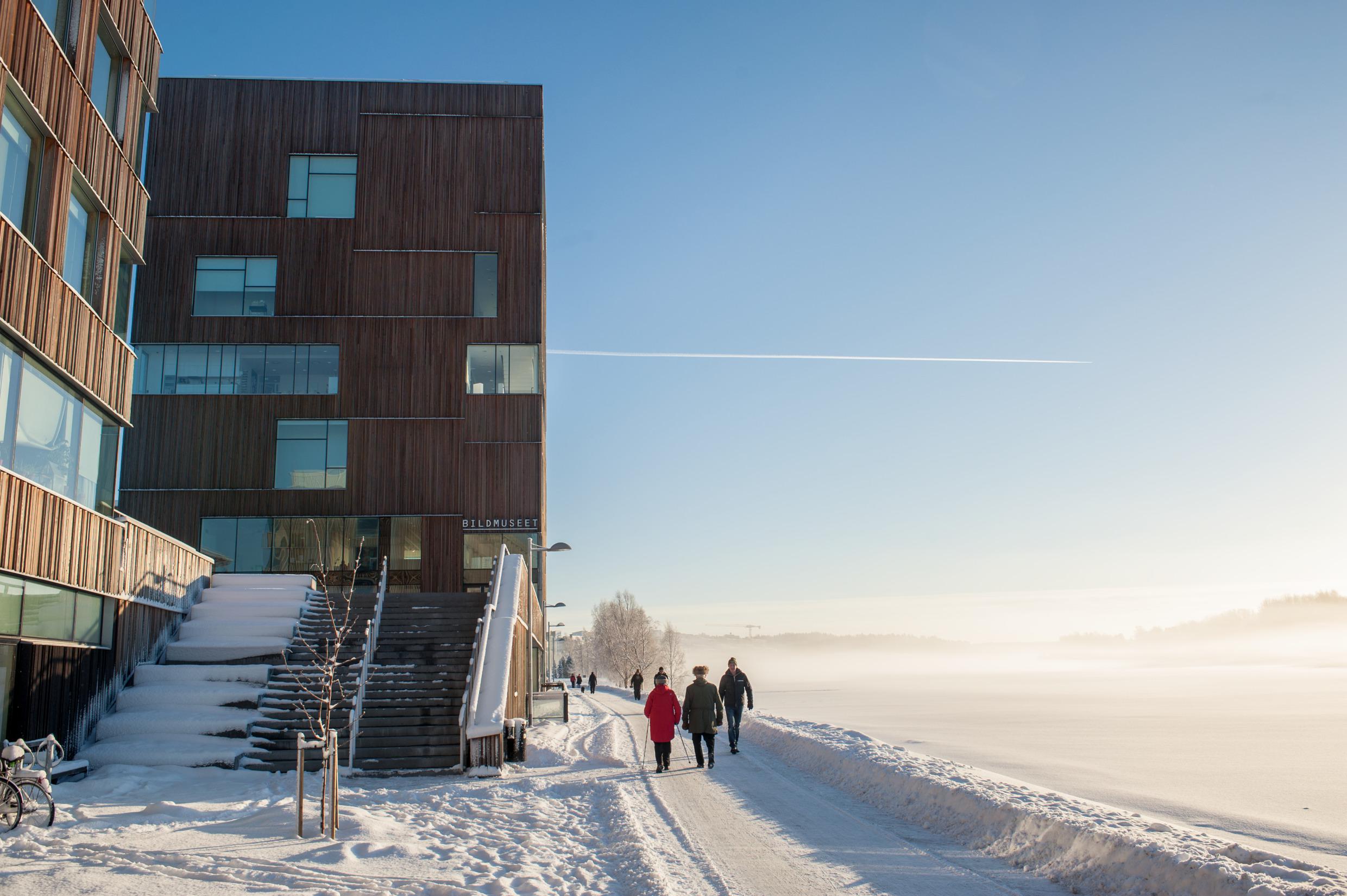 Links führt eine verschneite Treppe zu einem mehrstöckigen haus mit Holzwand und fenstern hinauf. Rechts gehen Leute auf einer von Schnee befreiten Straße neben einem Fluss,, der diesig von Sonne beschienen ist.