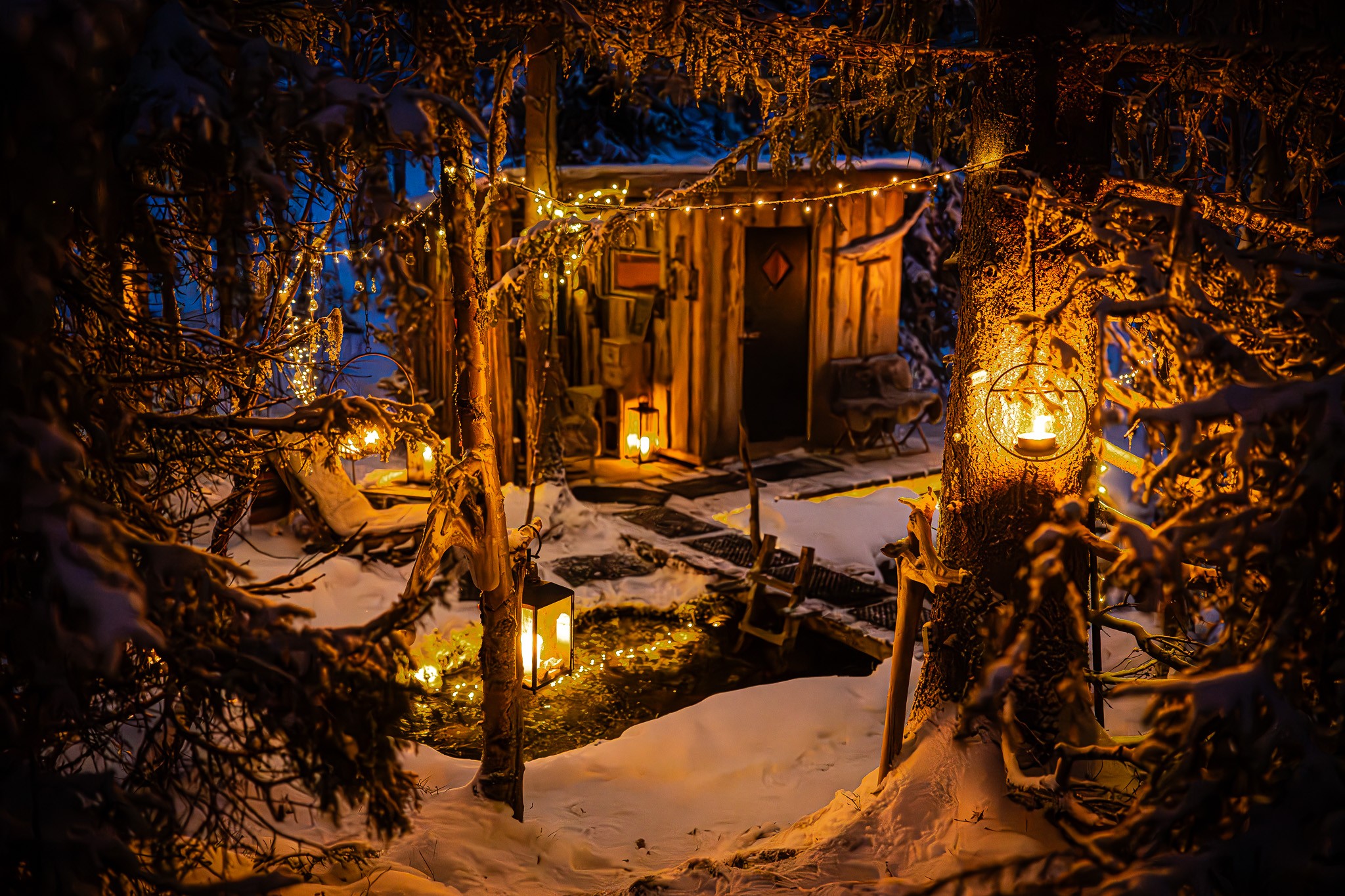 A small wooden sauna surrounded by snow-covered forest, lit by lanterns and string lights in the evening.