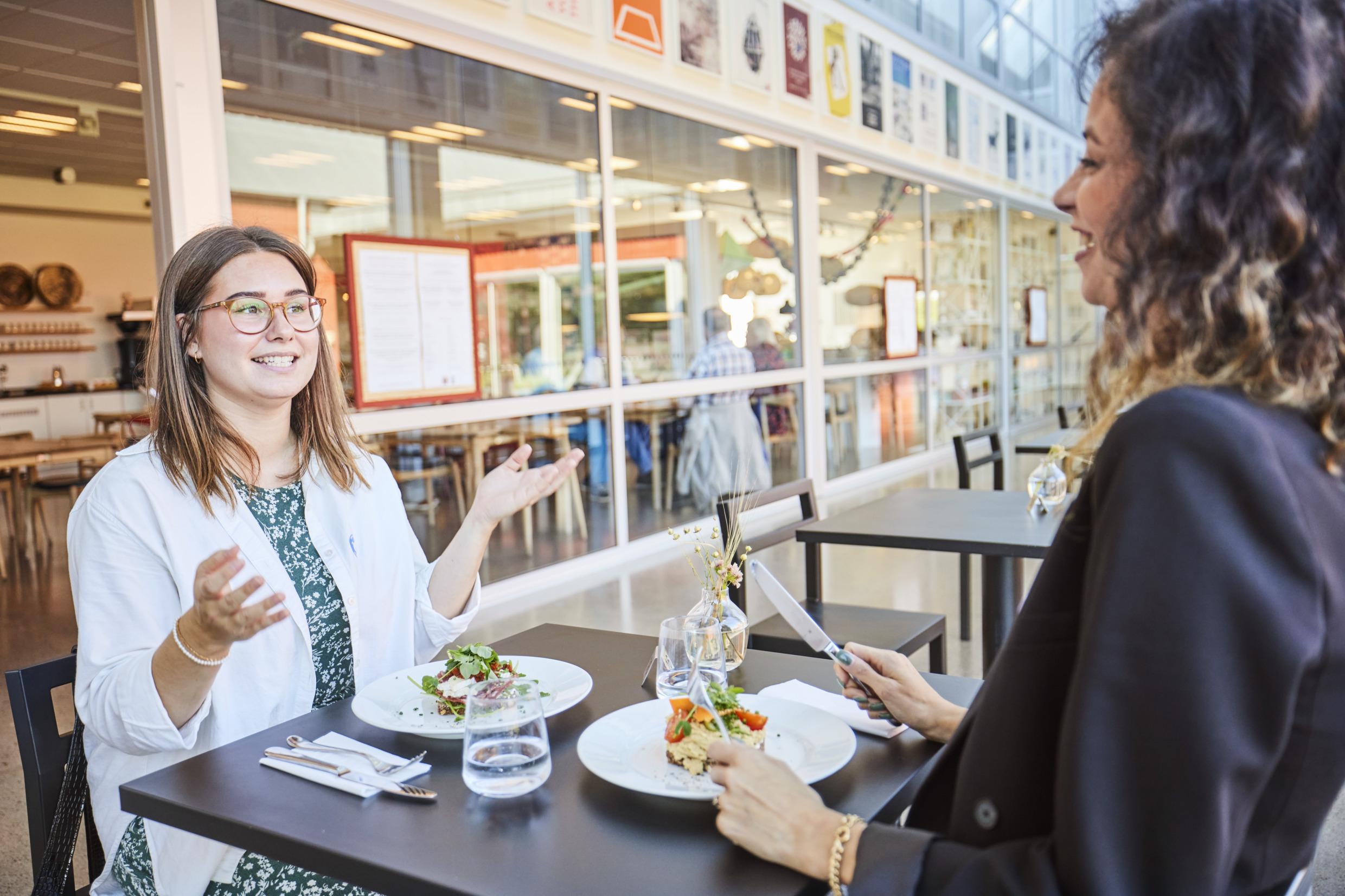 Twee vrouwen zitten aan een zwarte tafel te praten. Ze hebben borden met eten voor zich op tafel staan.