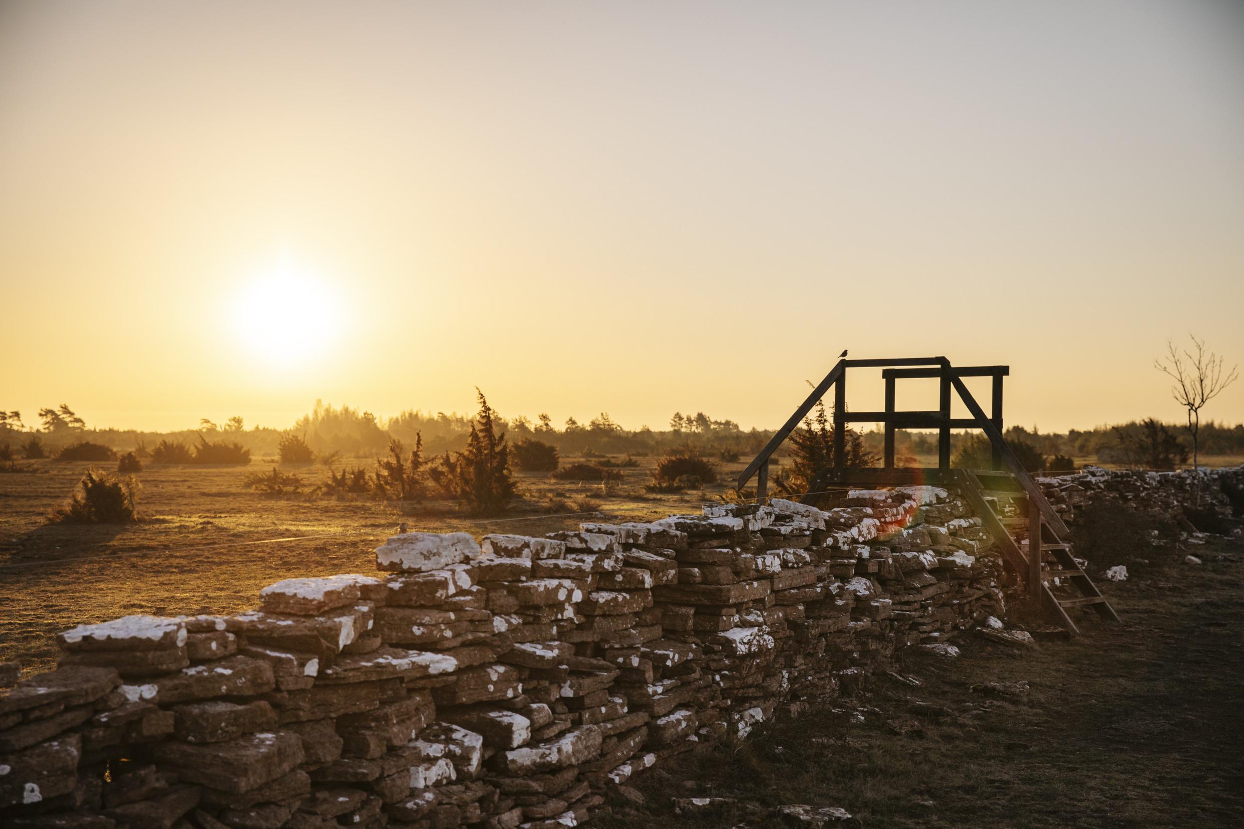 Eine Steinmauer, die teilweise mit Schnee bedeckt ist, die Sonne geht auf dem Feld dahinter unter.
