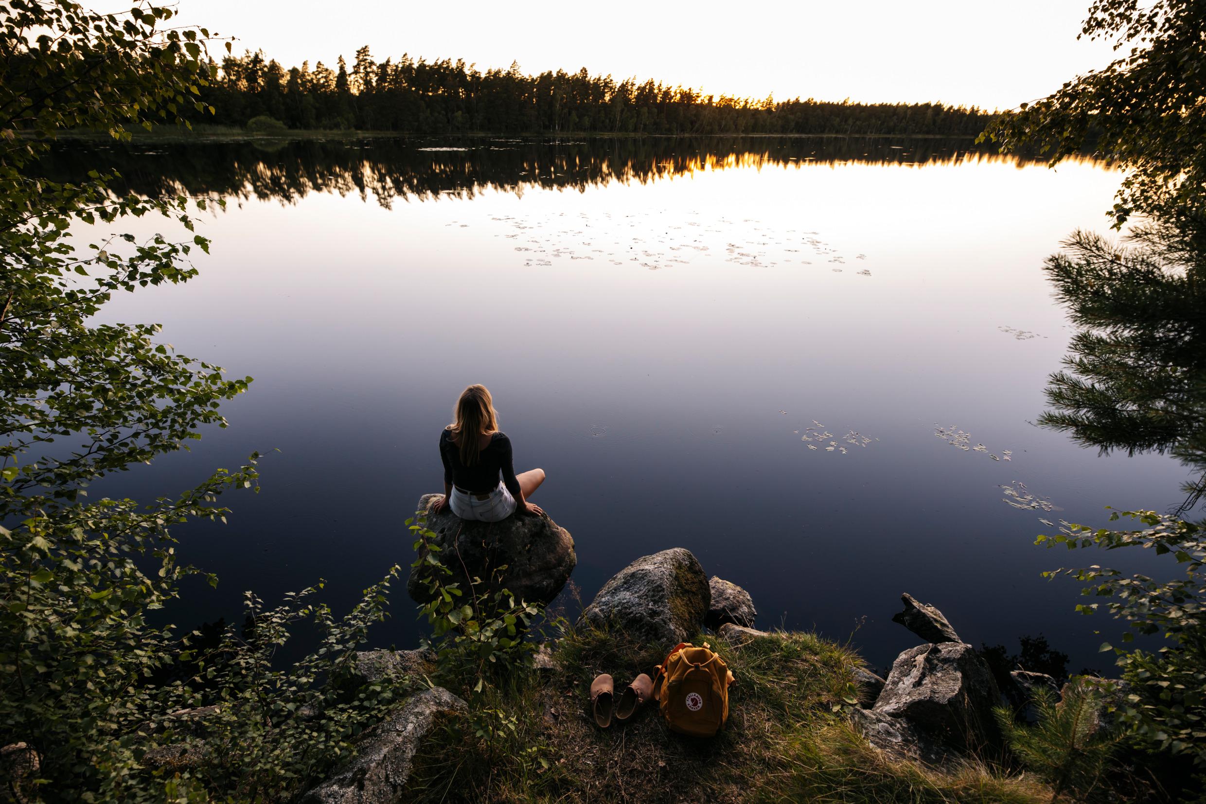 A woman sitting on a small rock looking out over a calm lake, the surrounding forest is mirrored in the lake.