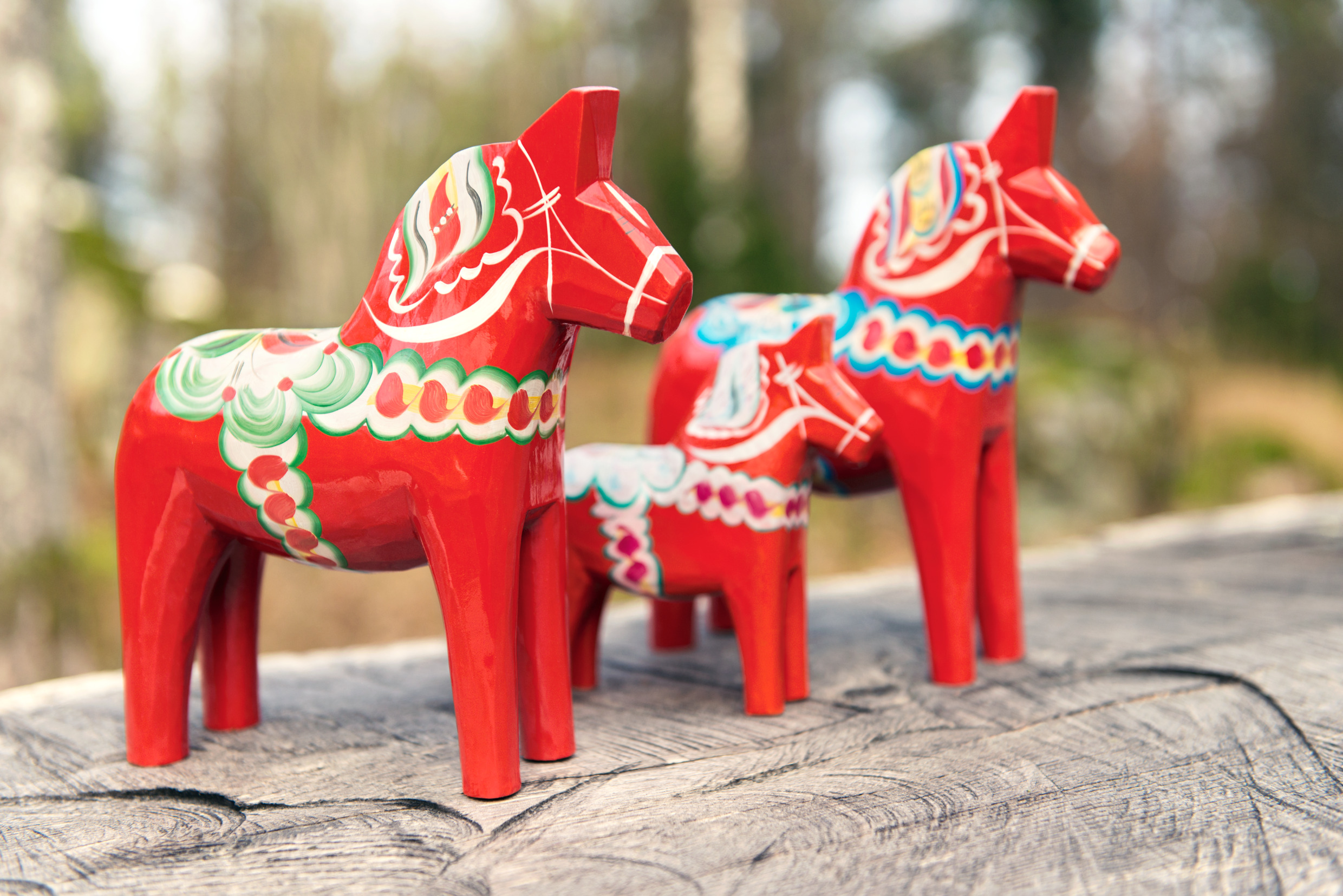 Three painted wooden Dala horses sits on a wooden rail with the Swedish forest in the background.