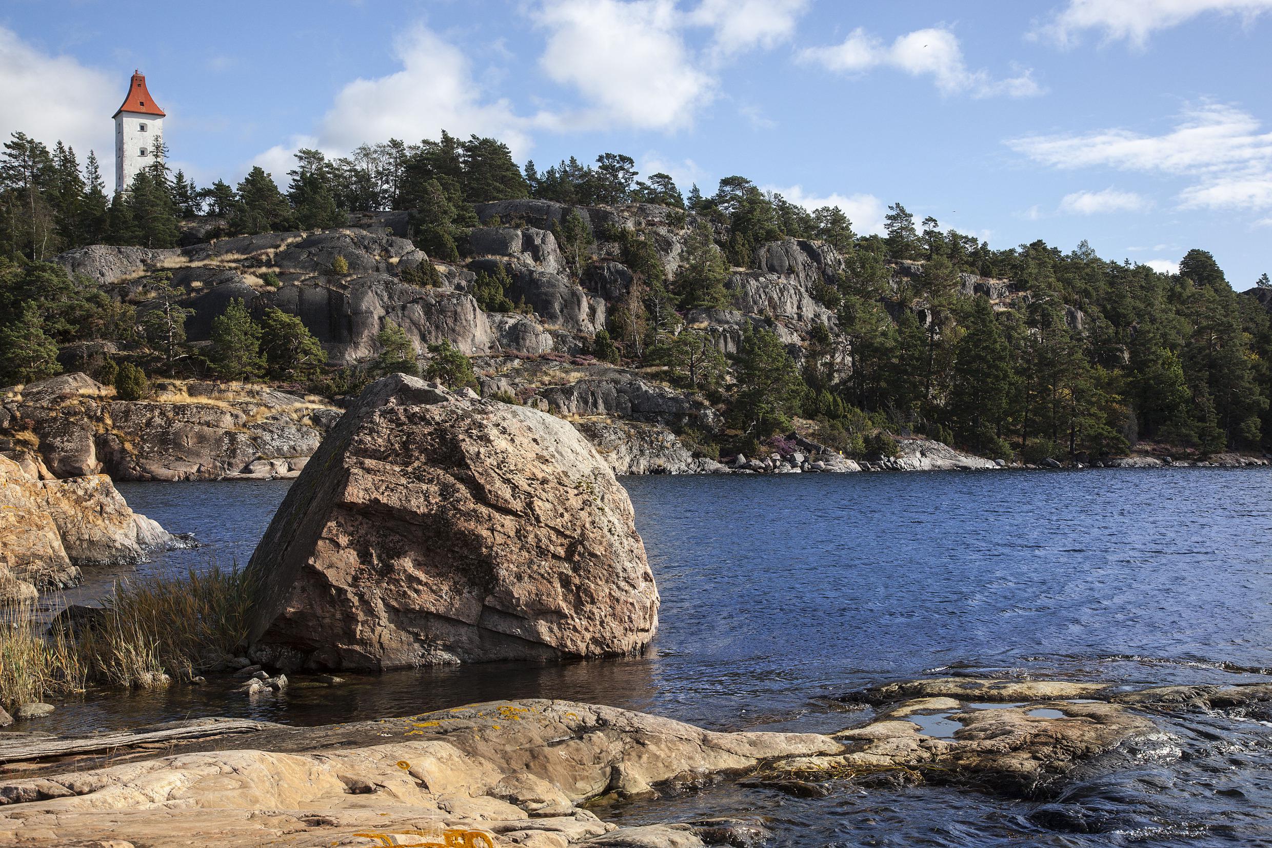 Des rochers et des arbres au bord de la mer, une tour blanche au loin.