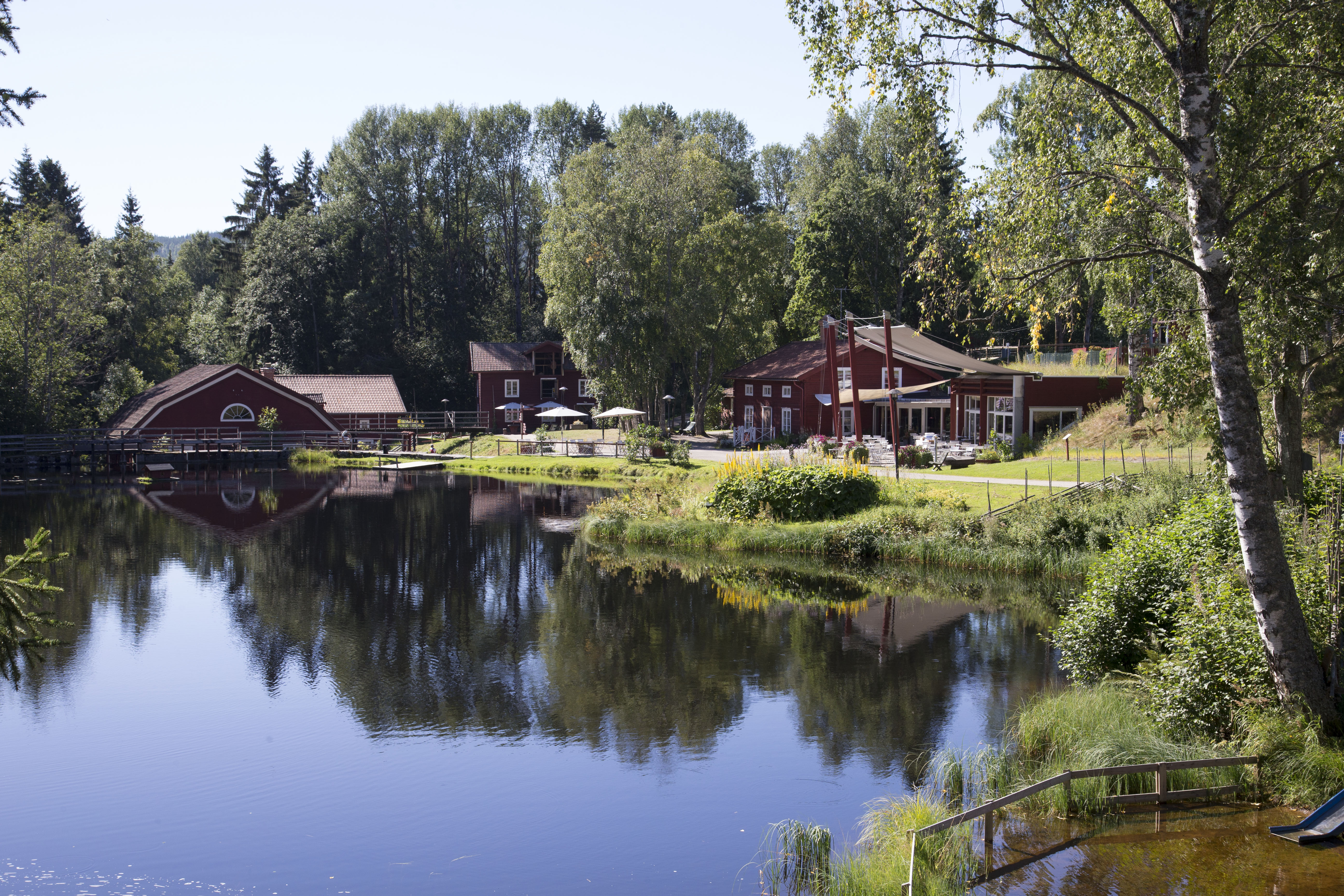 Red wooden buildings at Växbo Kvarn by a calm pond, surrounded by trees and greenery.
