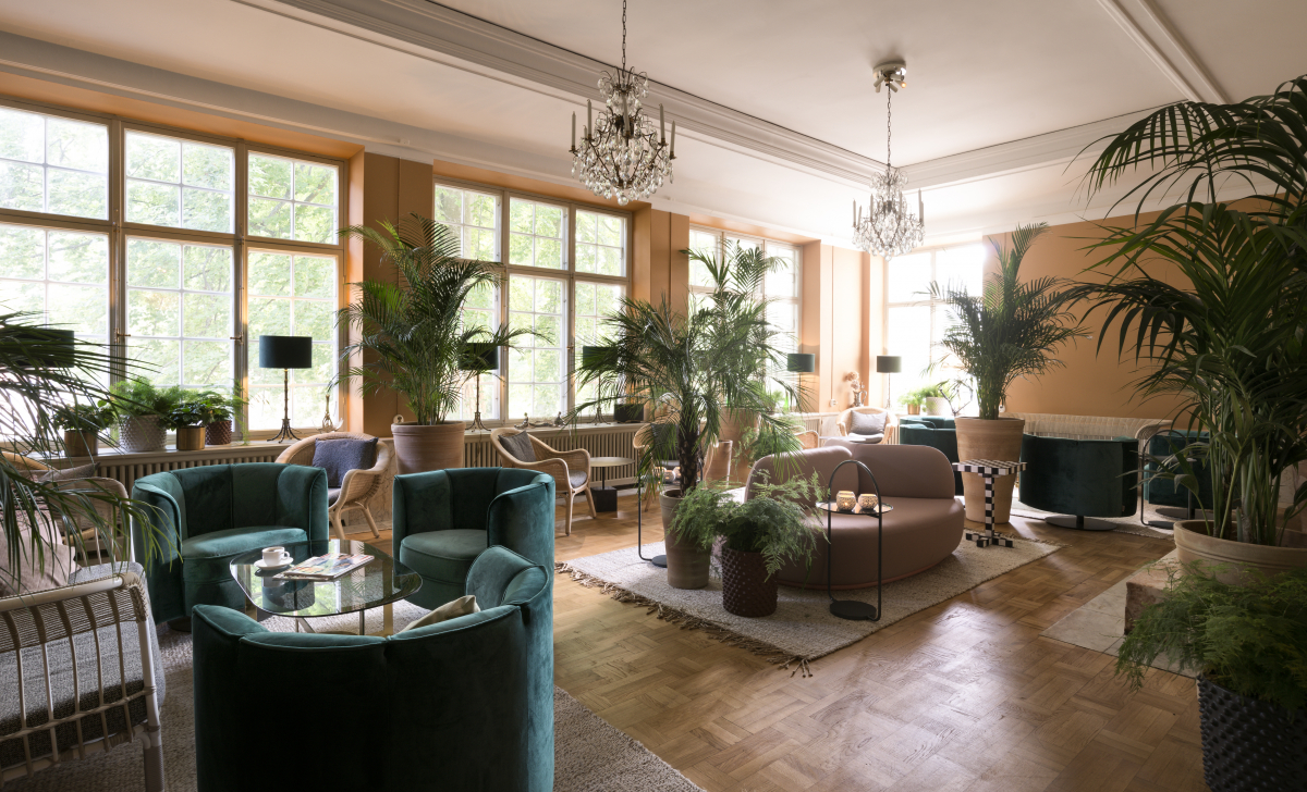 Un coin salon avec des canapés et des fauteuils. La pièce est décorée de plantes et de deux lustres suspendus au plafond.