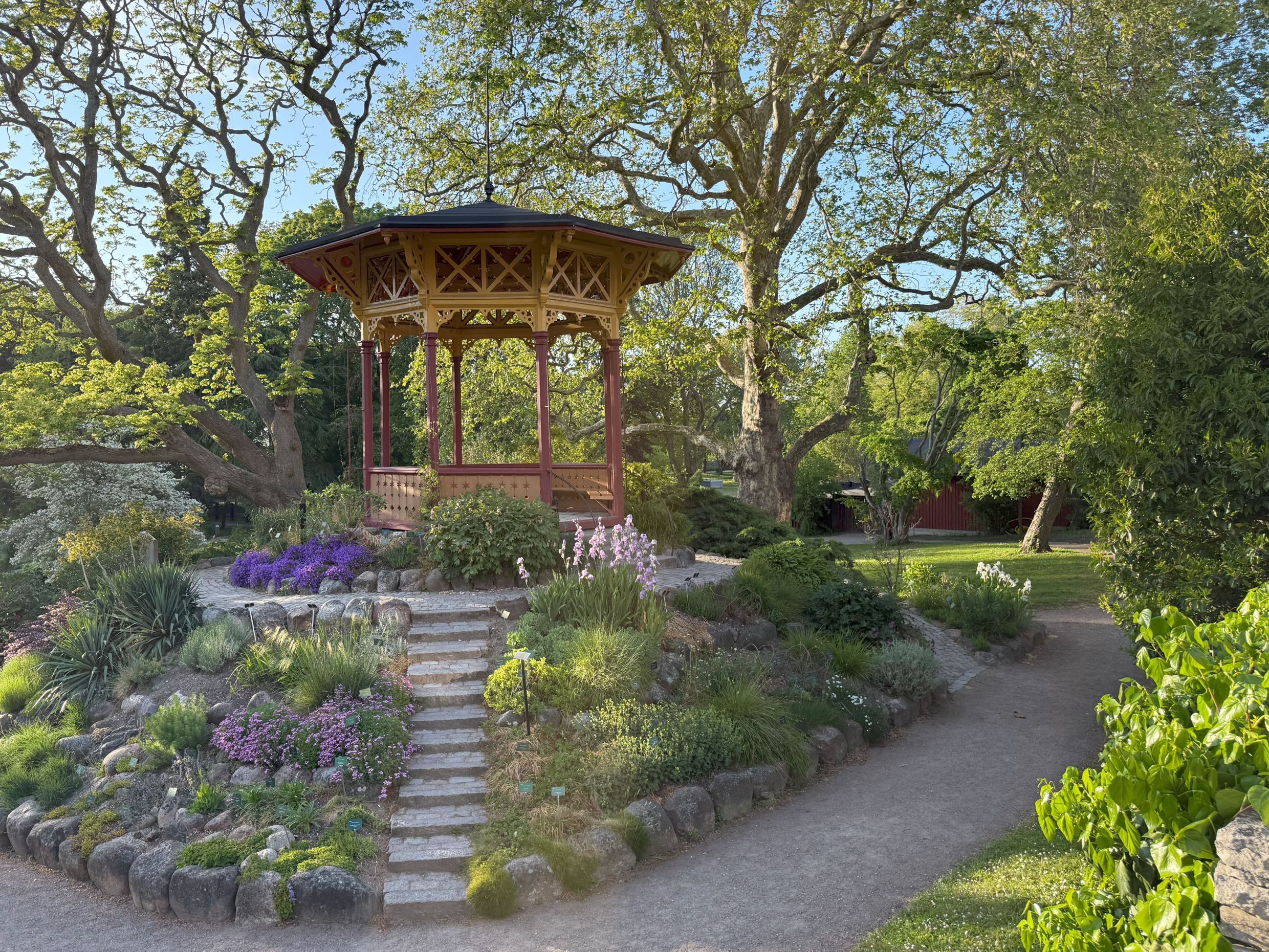 Wooden pavilion surrounded by lush greenery and exotic plants in Visby’s botanical garden on a sunny summer day.