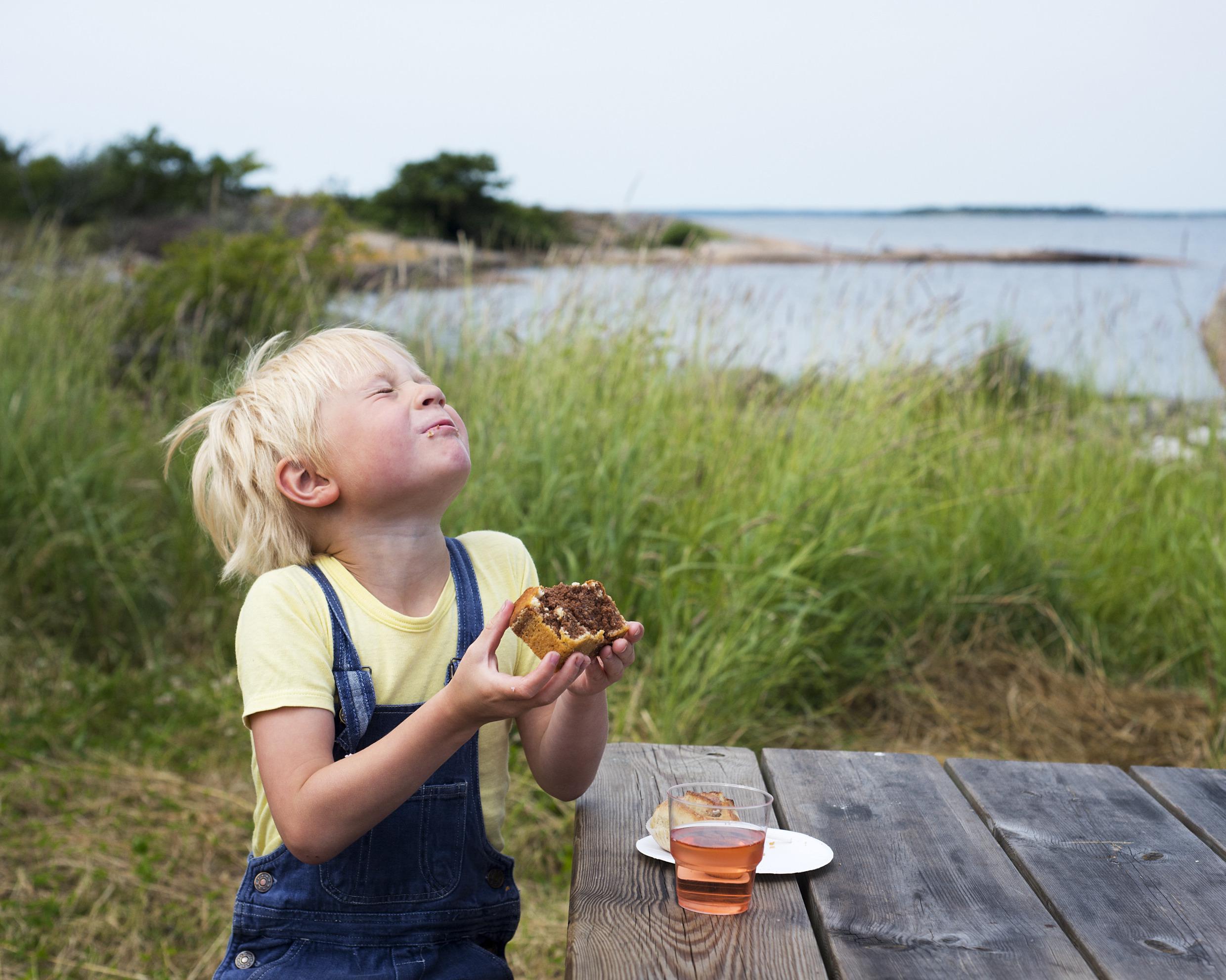 Een jongen zit op een bankje, drinkt een sapje en eet een stukje cake.