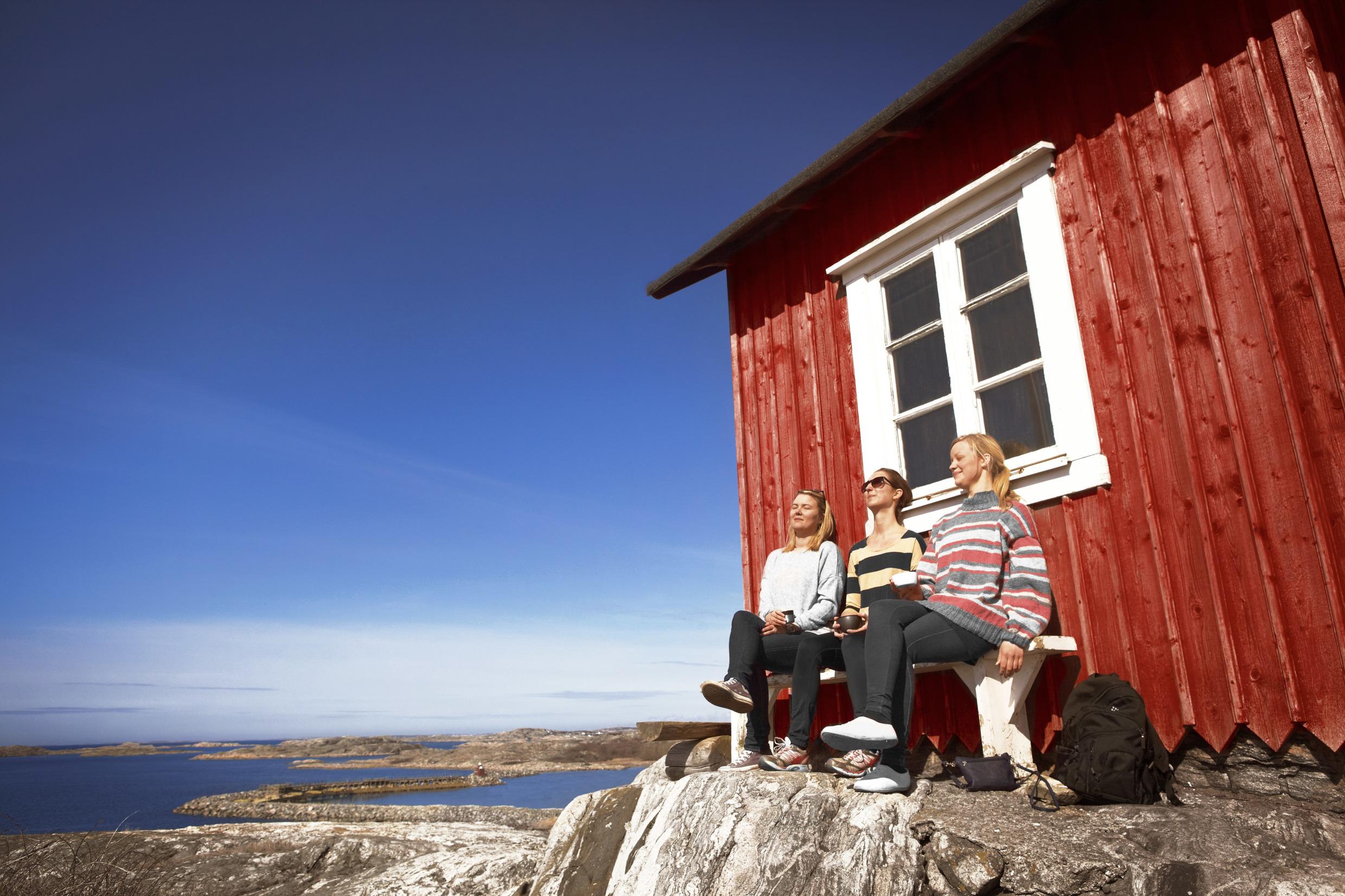 Three women resting against a red wooden house, on a rocky island. The sky is blue and the sun is shining.