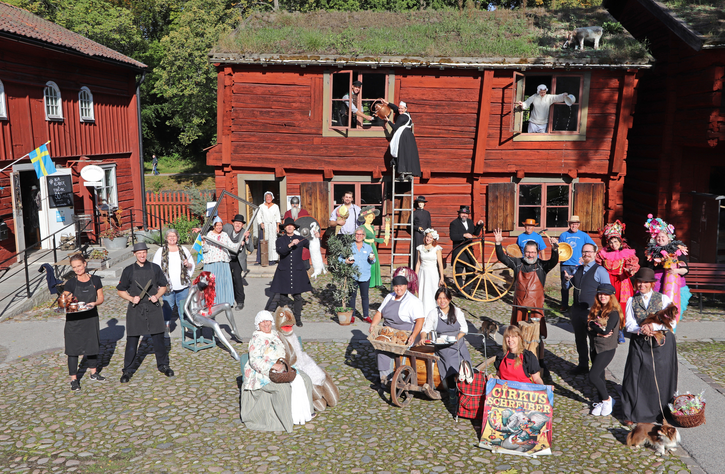 Een heleboel mensen in historische en theaterkostuums stonden bij elkaar voor rode houten gebouwen in het openluchtmuseum Wadköping in Örebro, Zweden.