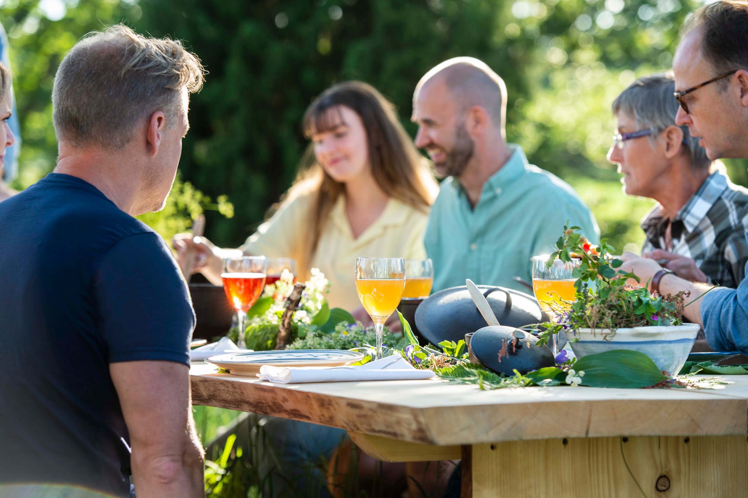 Five persons sitting around a wooden table set with plates and wine glasses. There is a cooking pot and fresh herbs on the table.