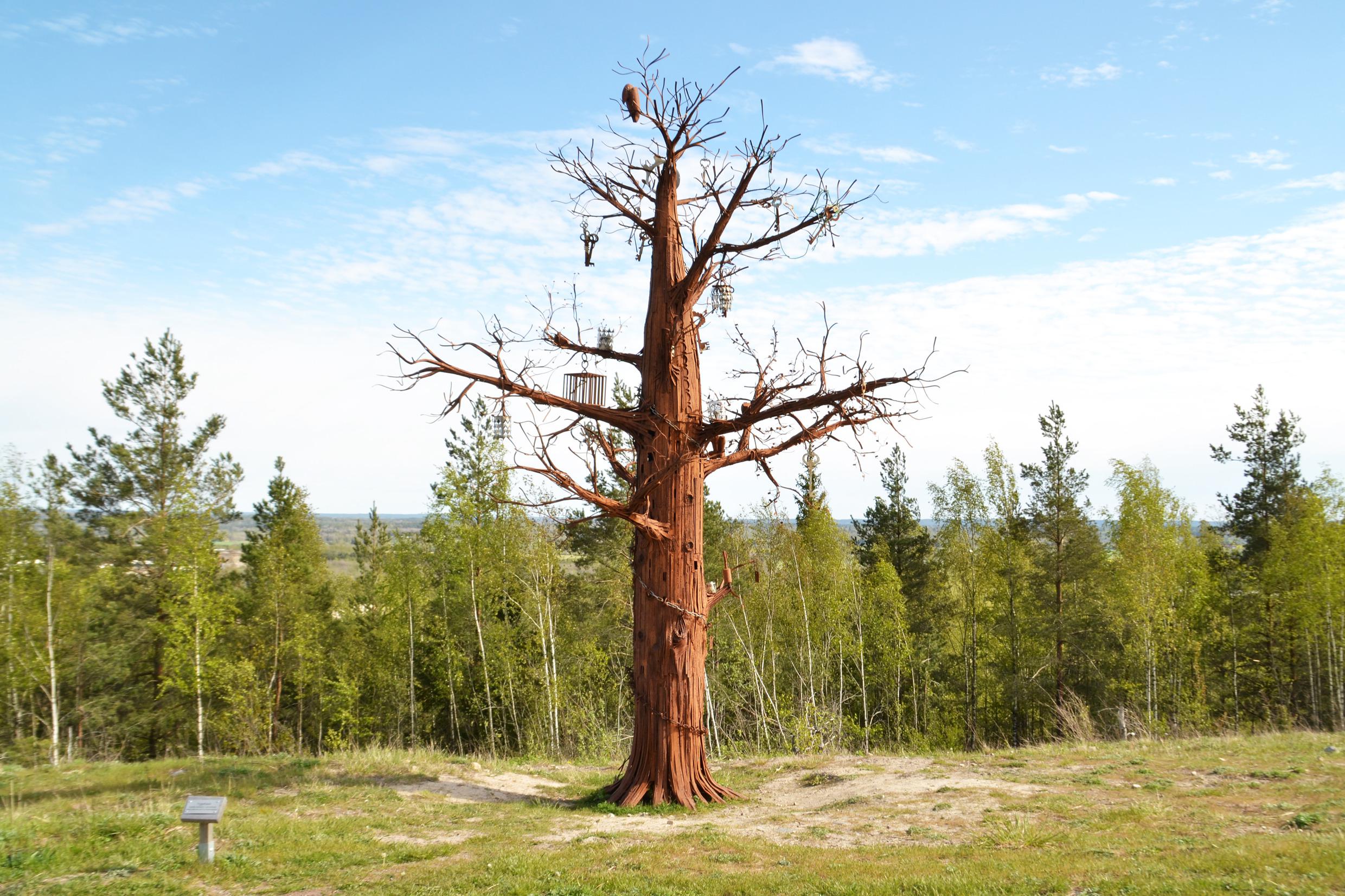 Eine Skulptur namens „Yggdrasil“. Ein Baum aus Metall, der mit vielen Schlüsseln und anderen Gegenständen geschmückt ist, steht auf einem Hügel in einem Skulpturenpark mit einem Wald im Hintergrund.