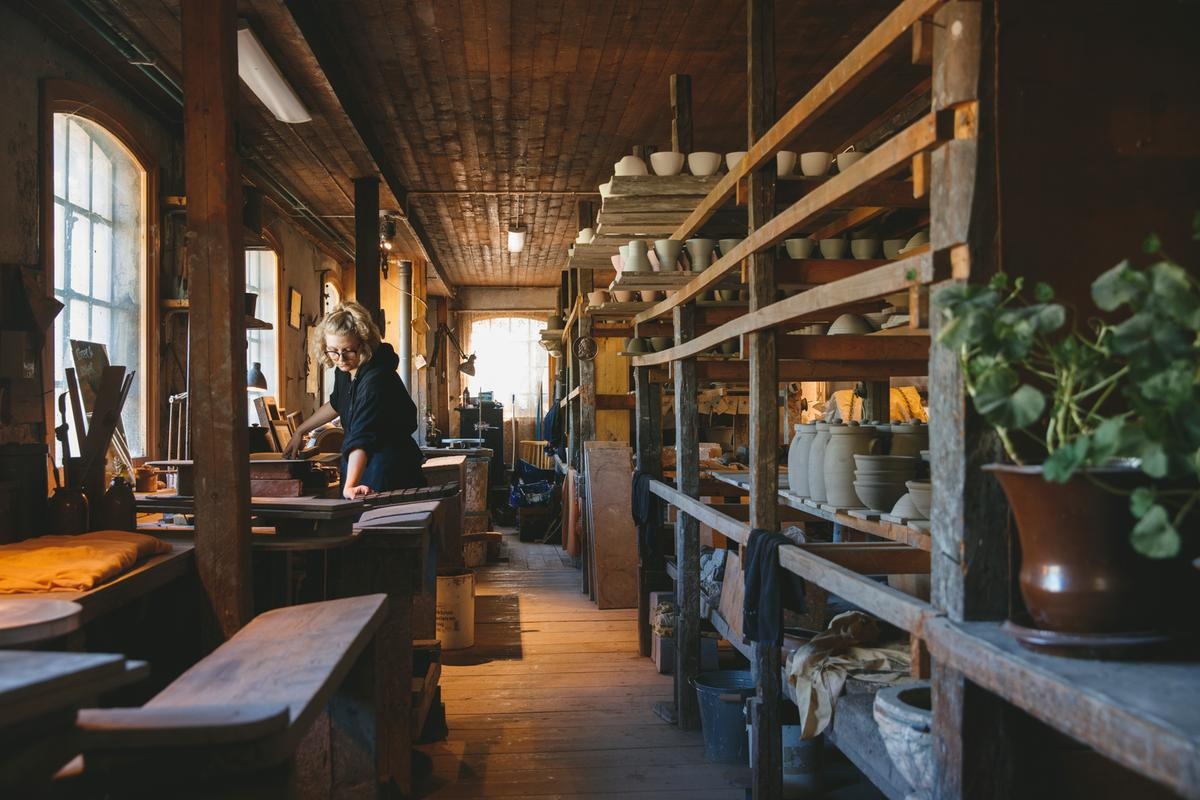 Potter working by hand in the rustic workshop at Wallåkra Stoneware Factory, surrounded by shelves of handmade ceramics.